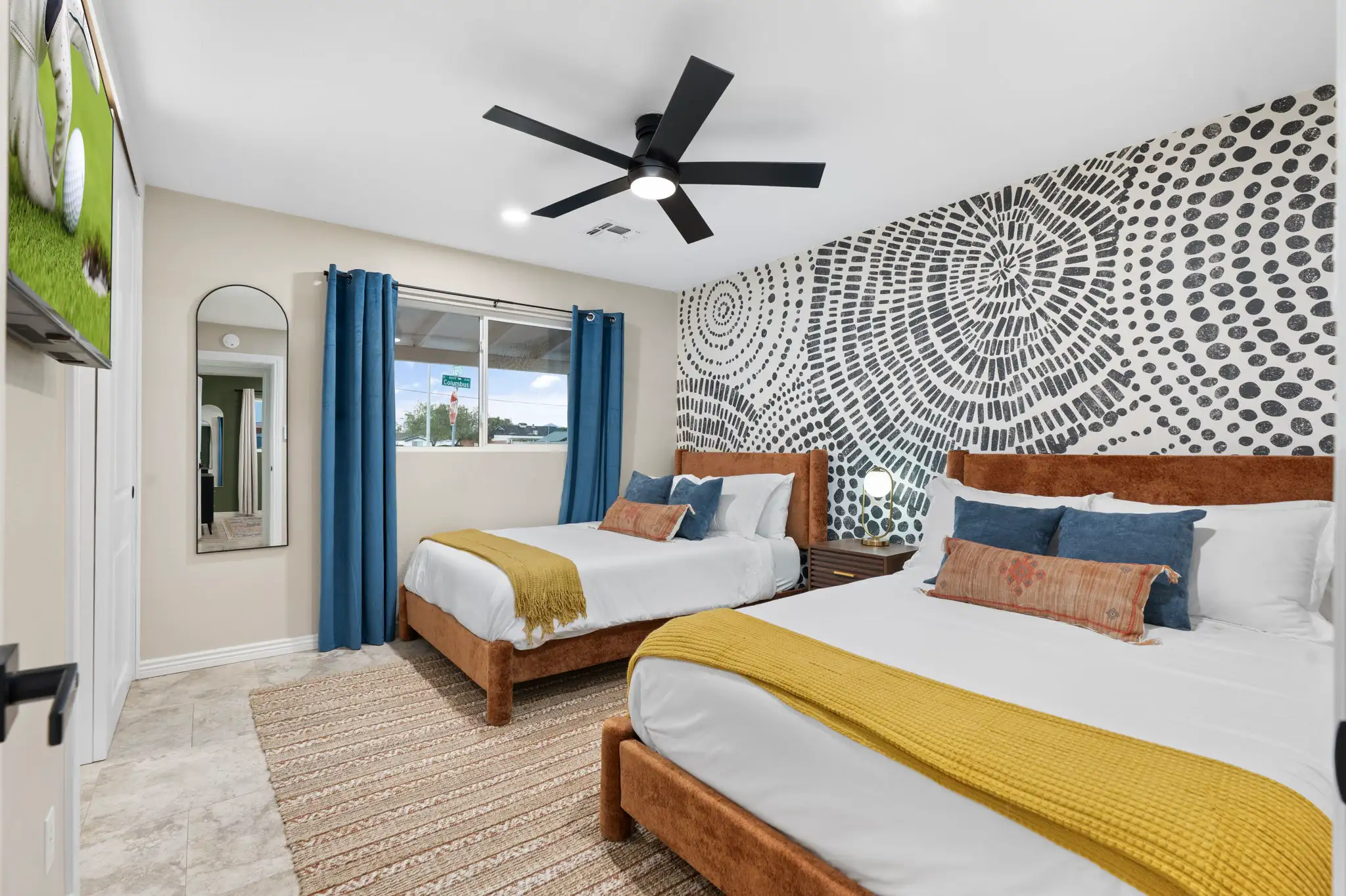 A modern bedroom with two beds, featuring rust-orange headboards and yellow throws. A bold, black and white geometric spiral pattern covers the accent wall.