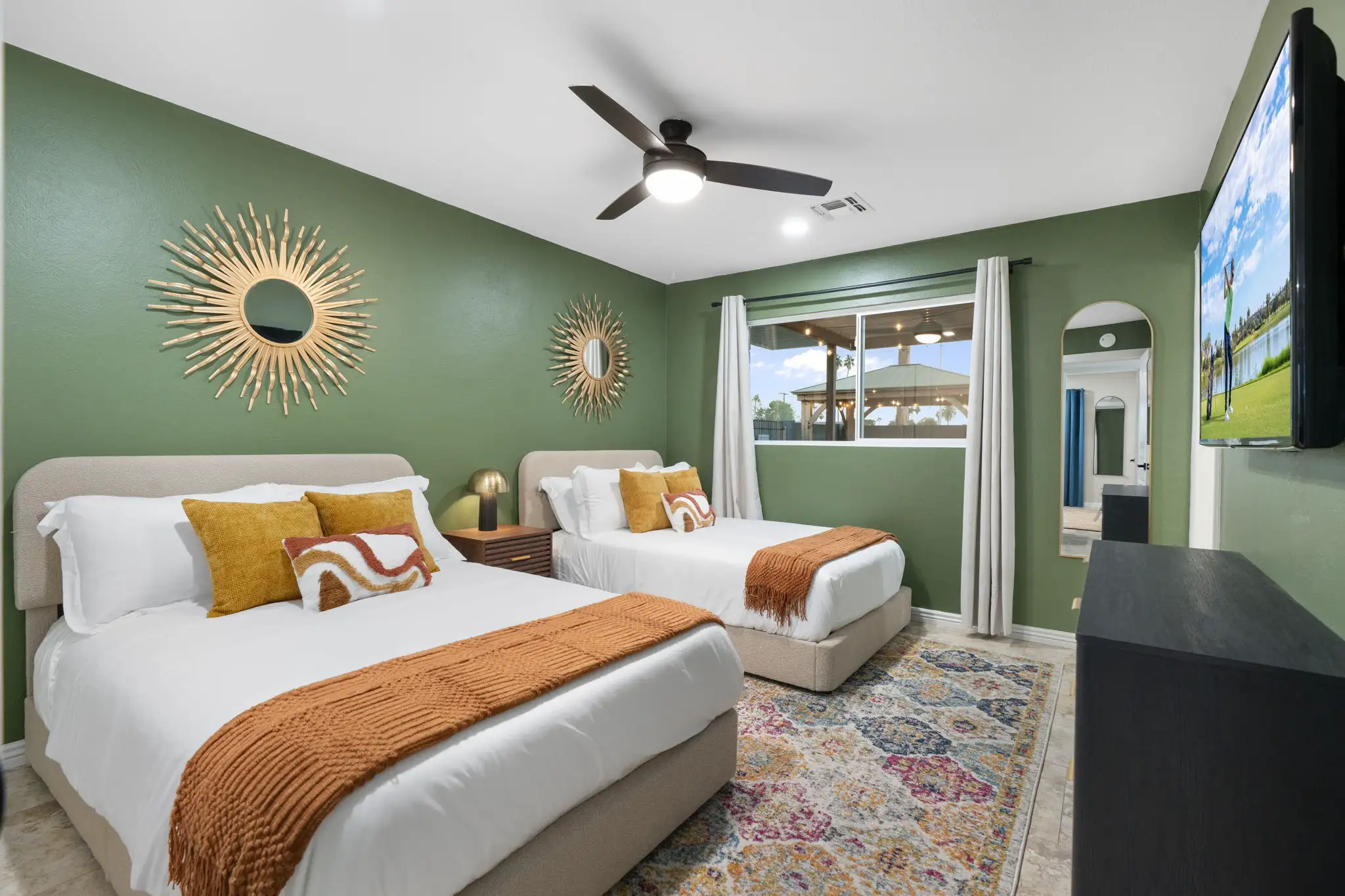 A bright bedroom featuring two beds with white linens and rust-colored throws, set against olive green walls decorated with sunburst mirrors and a colorful rug.