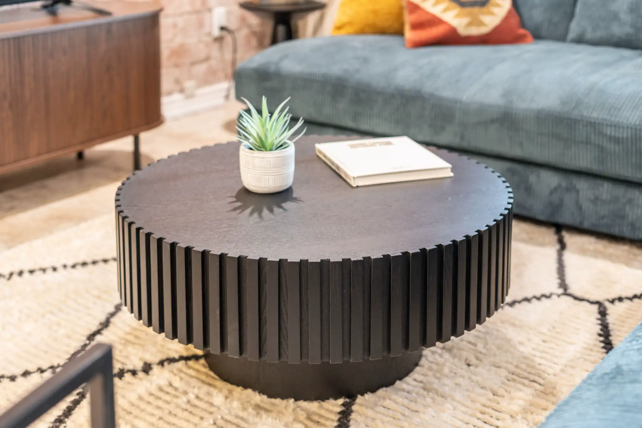 A dark, ribbed circular coffee table sits on a geometric rug. The table holds a small potted succulent and a white book, complementing the blue corduroy sofa.