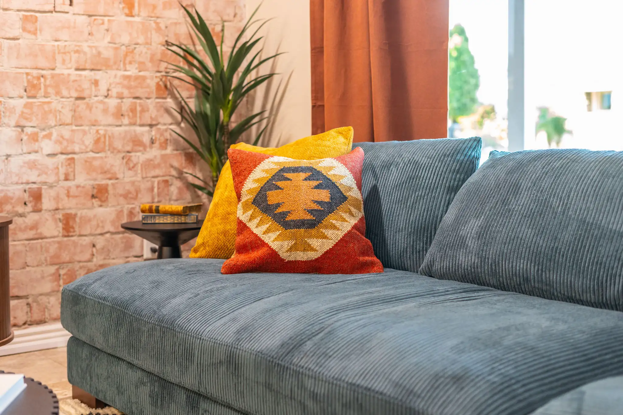 A blue ribbed sofa is set against an exposed brick wall. It holds two accent pillows: one solid yellow and one featuring a striking red, orange, and blue geometric pattern.