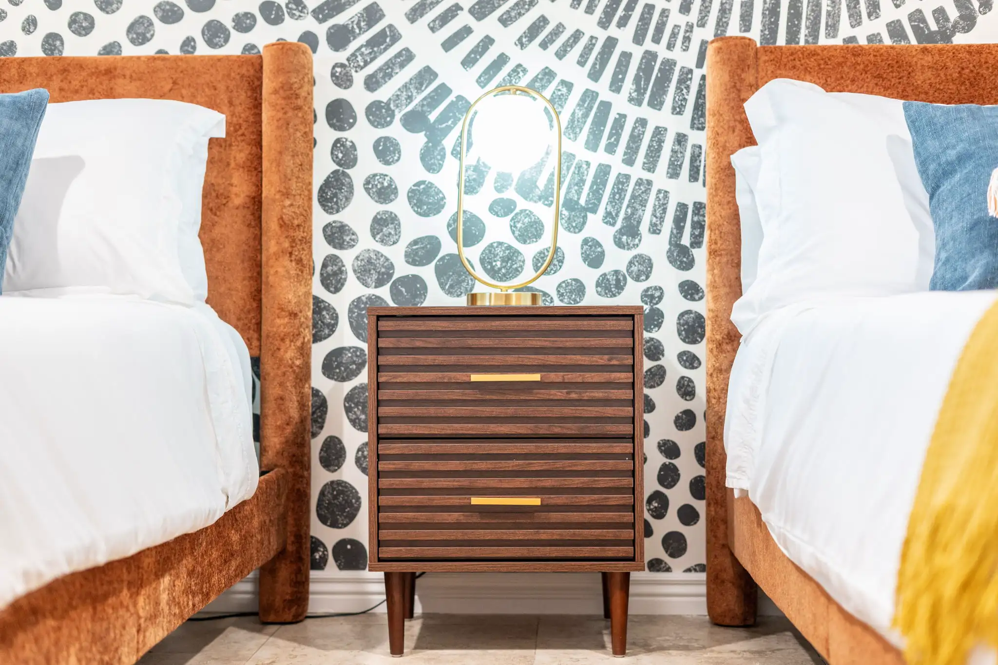 A dark wood nightstand with a brass lamp is centered between two beds with tall, orange velvet headboards. The wall behind features bold abstract wallpaper.