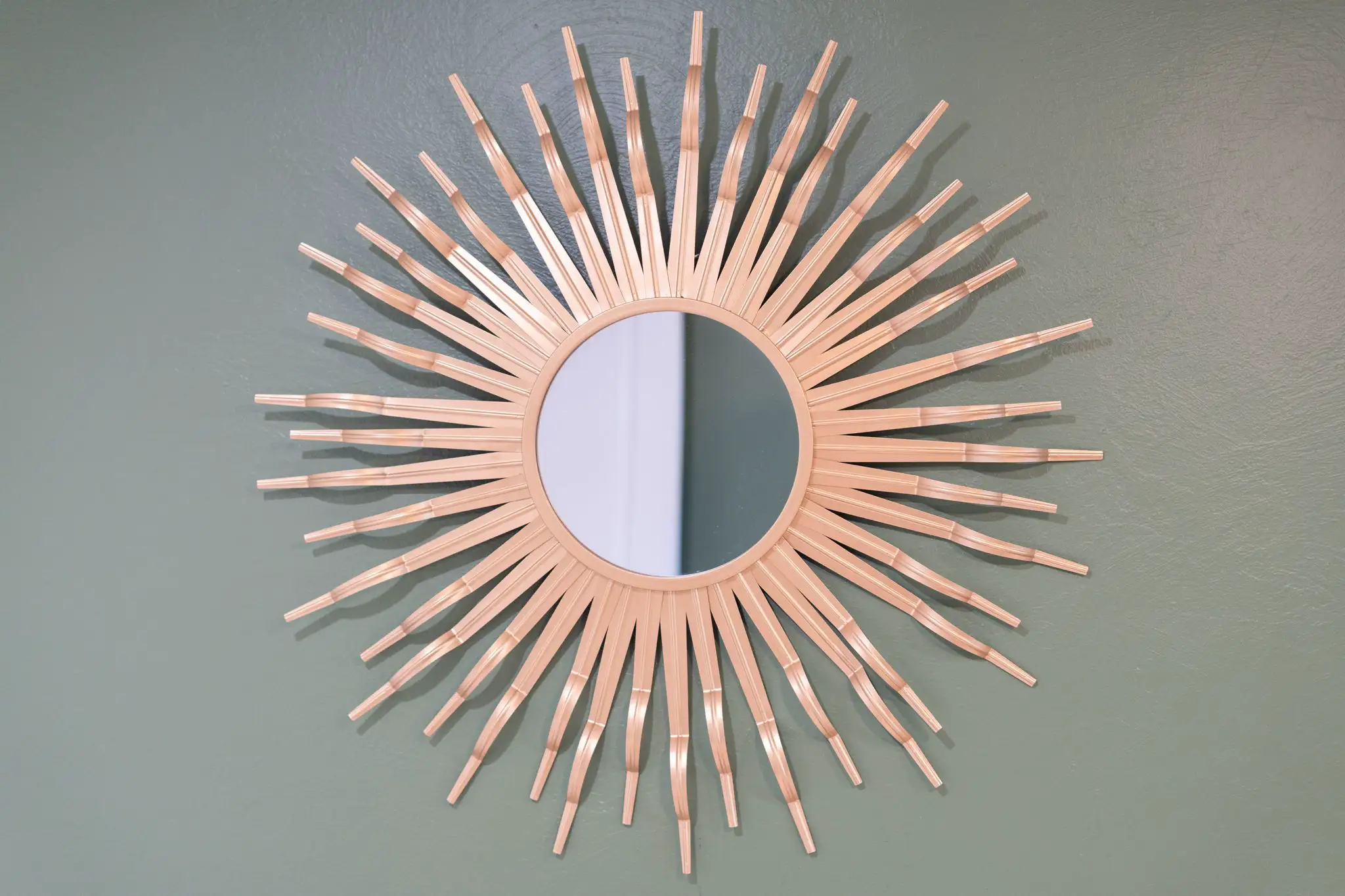 A round sunburst mirror with a natural bamboo frame is mounted on a wall painted a muted green color in a rental property.