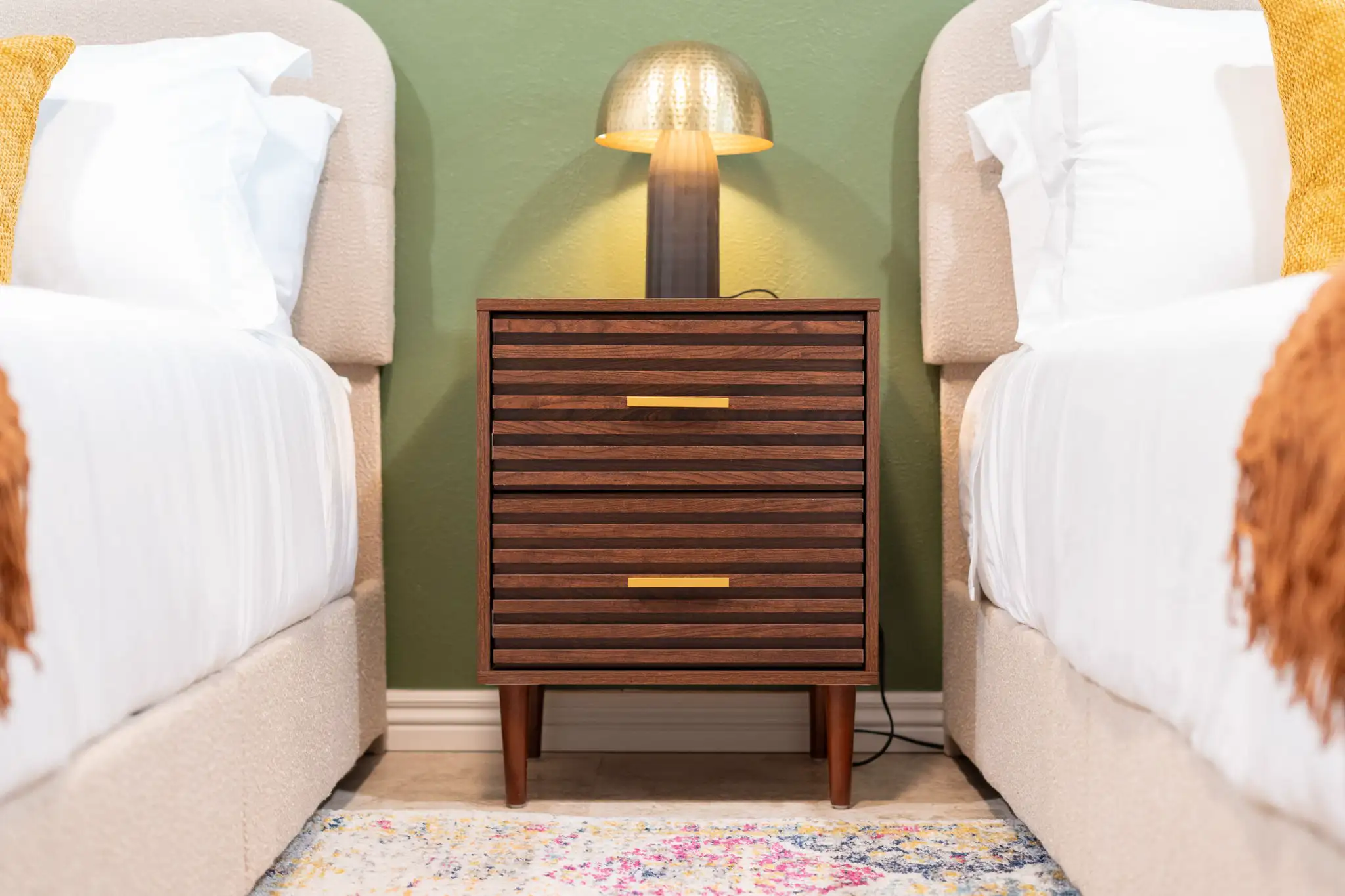 A symmetrical bedroom view featuring a mid-century wood nightstand with gold handles, centered between two white beds. A warm brass lamp lights the sage green wall.