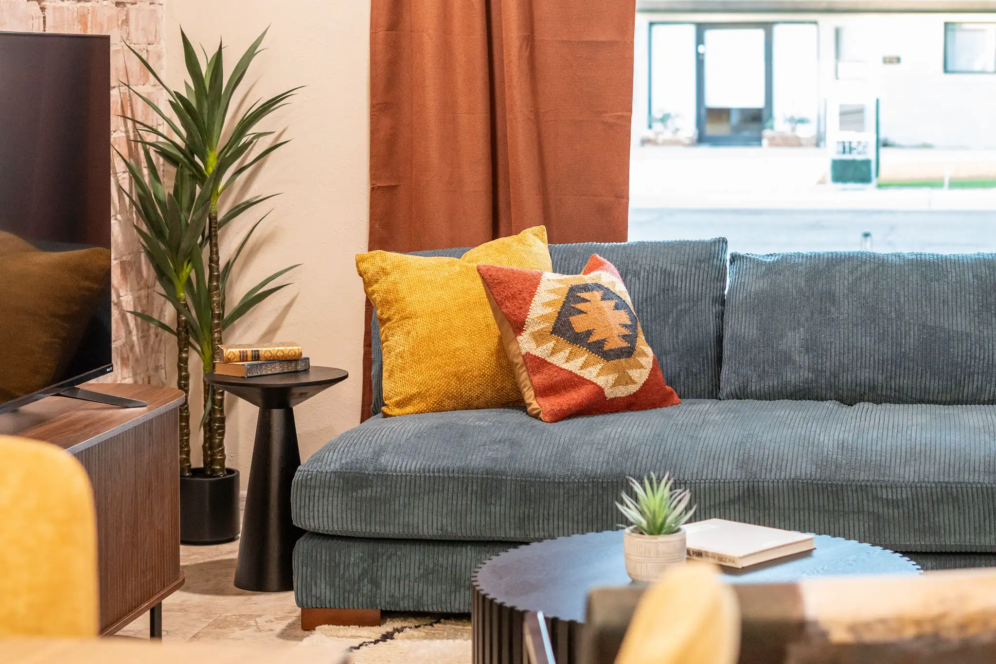 A comfortable seating area featuring a plush blue corduroy sofa and patterned pillows, next to a window with warm rust-colored drapes.