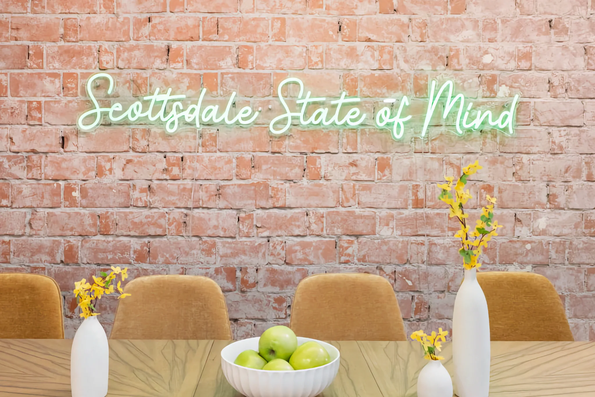 A mint green neon sign spelling "Scottsdale State of Mind" is set against a brick wall. A table below features green apples and yellow flowers.
