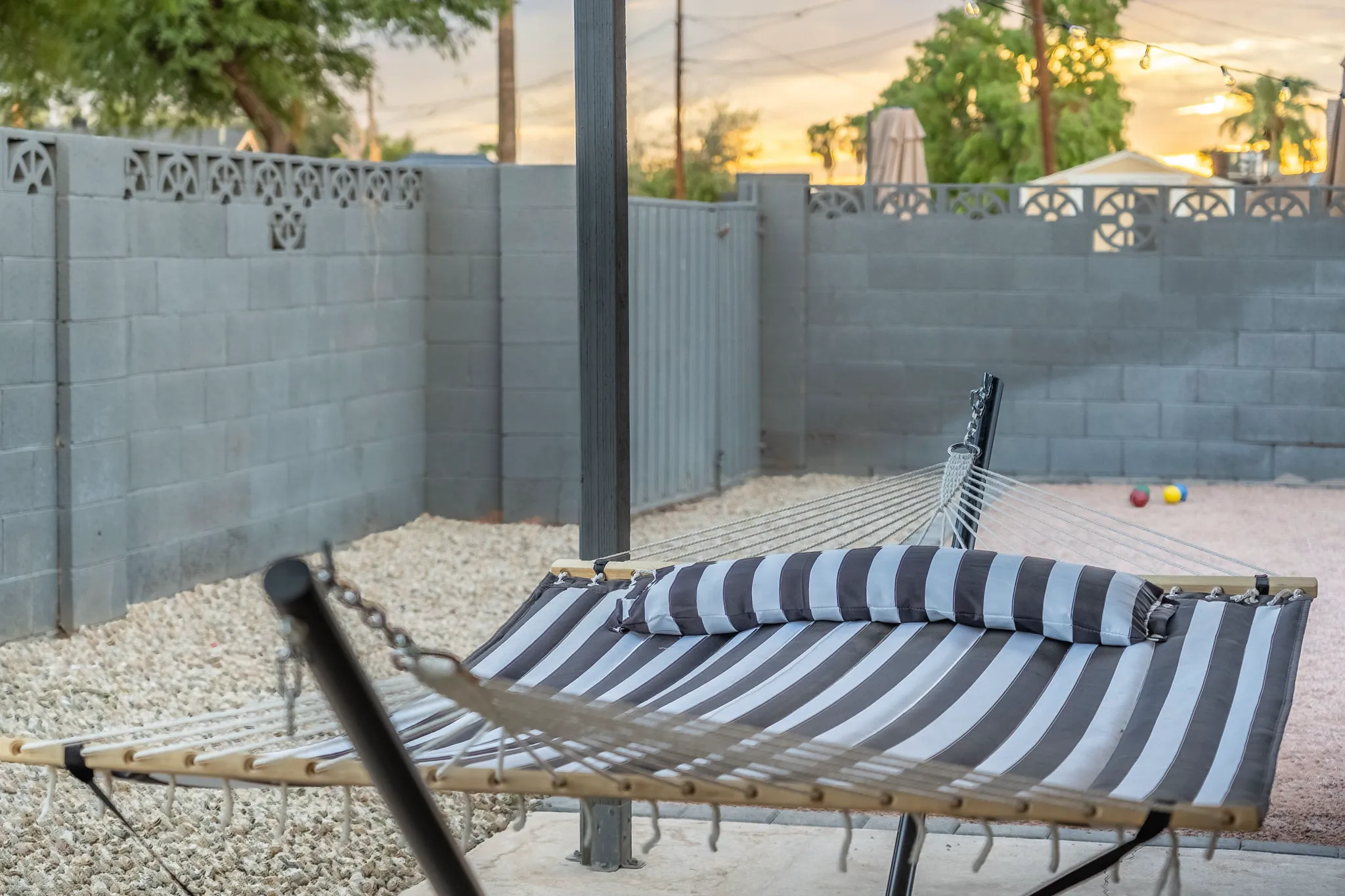 A black and white striped hammock with a matching bolster pillow sits on a gravel patio, enclosed by a gray concrete block wall, under string lights.