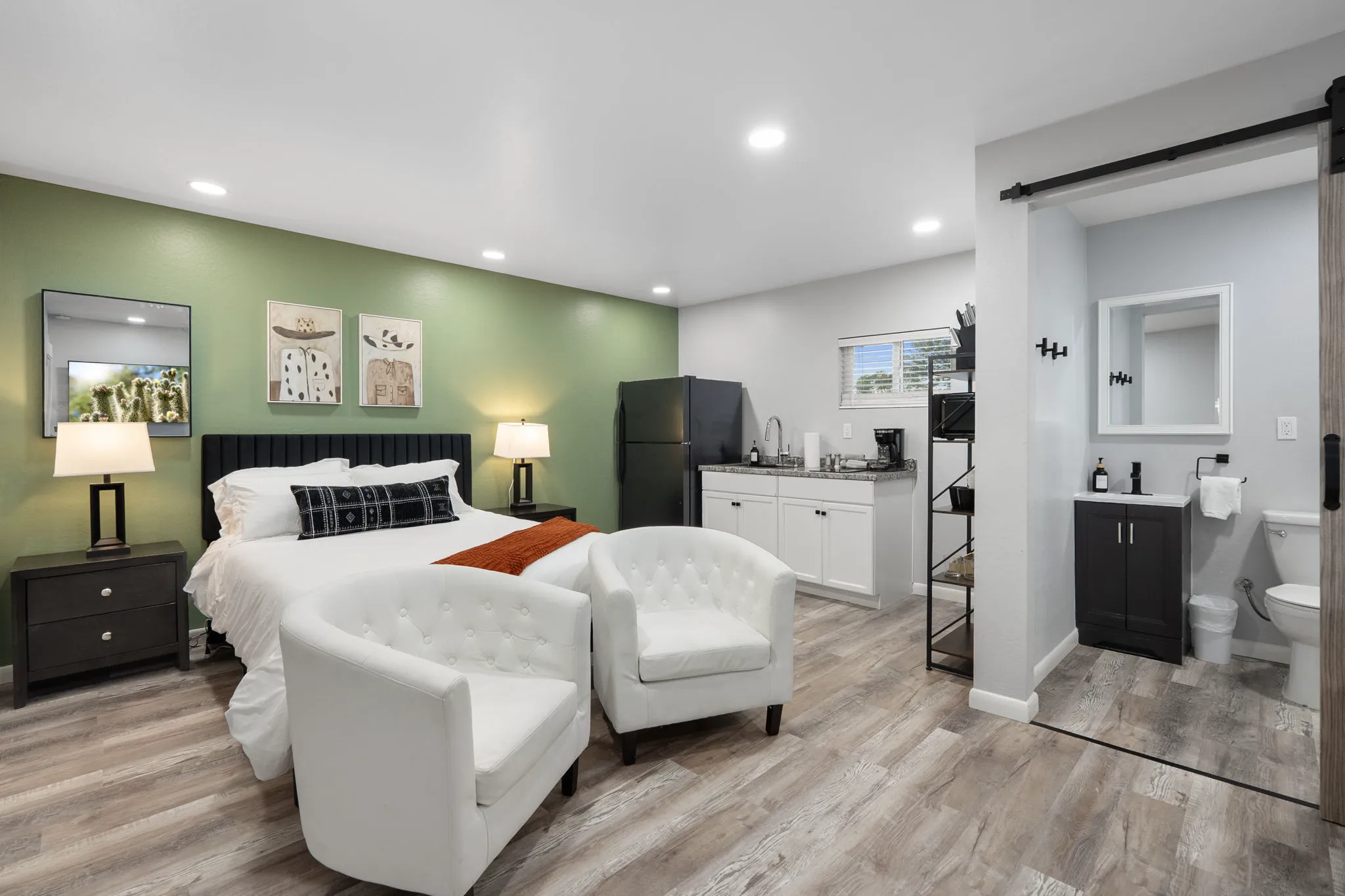 A studio apartment features a bed with an orange throw, green accent wall, and two white armchairs. A kitchenette with a black refrigerator and a partial bathroom are visible.