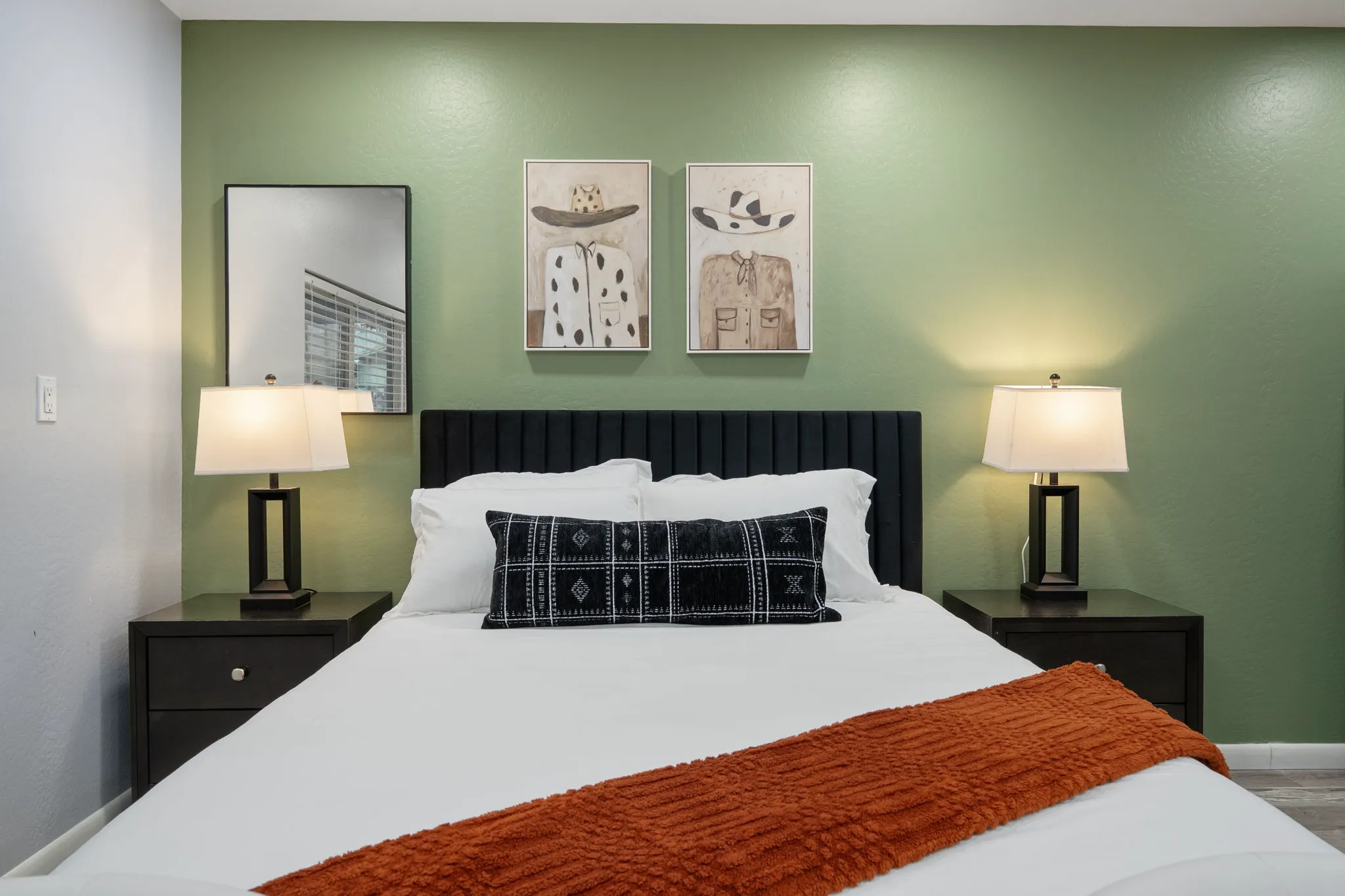 A neatly made bedroom features a black headboard, white bedding, an orange throw, and a patterned black pillow. Two black nightstands hold lamps, and framed hat-and-shirt art hangs on the green wall.