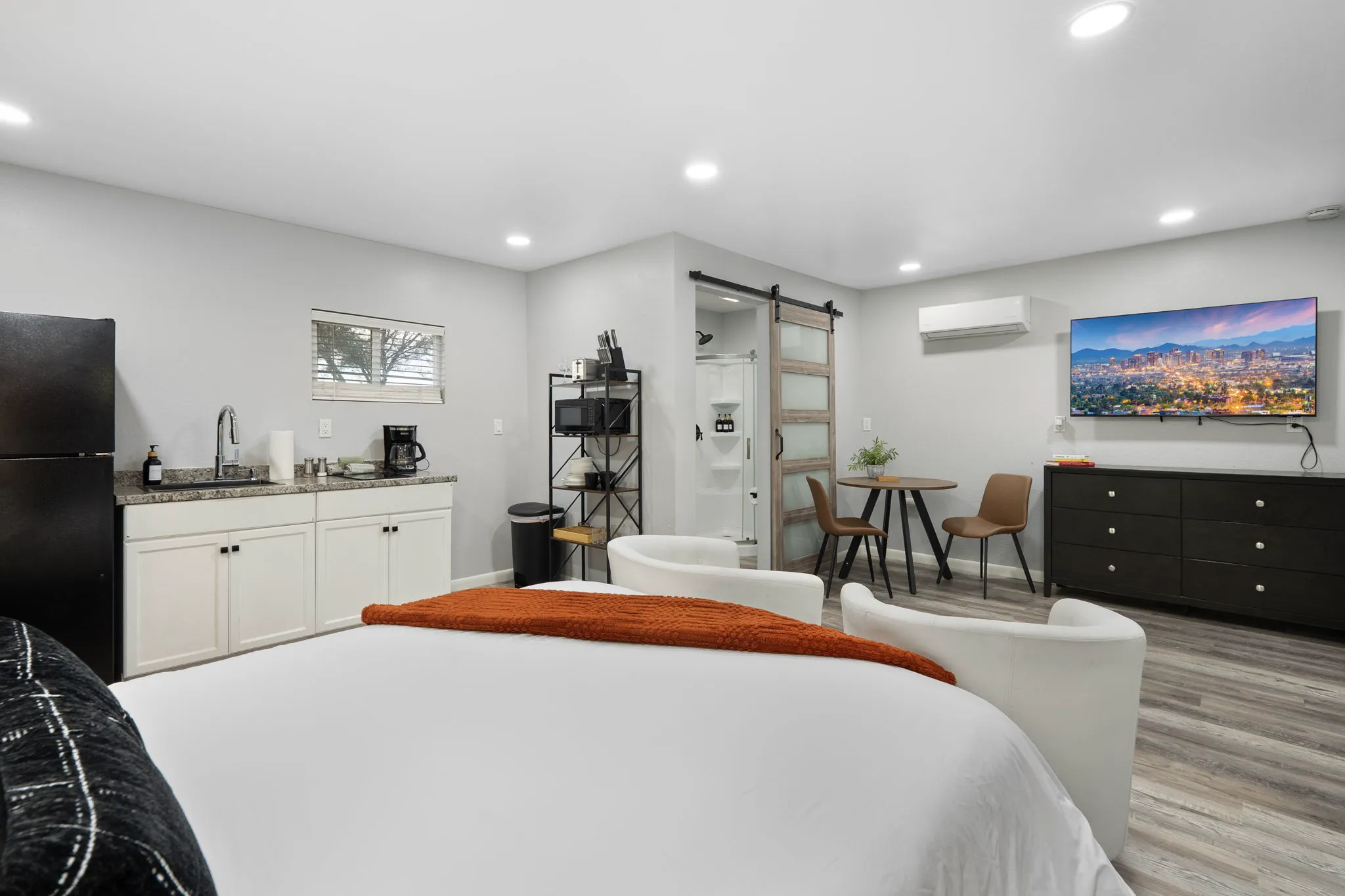 An open-concept studio features a white bed, a kitchenette, a small dining table with two chairs, and a TV mounted above a dresser. A barn door leads to a shower.
