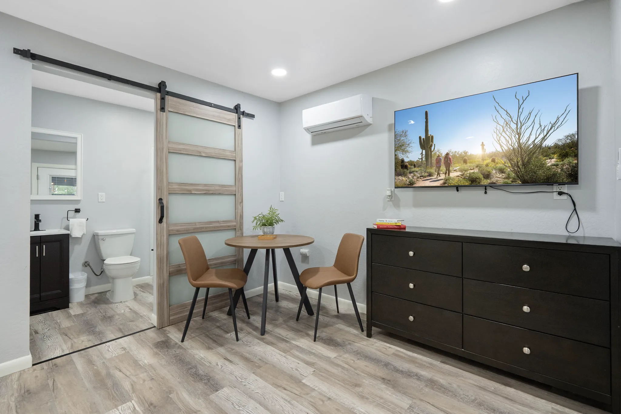 A modern vacation rental room with light wood floors. It features a TV above a dark dresser, a small round table with two brown chairs, and a sliding barn door leading to a bathroom.