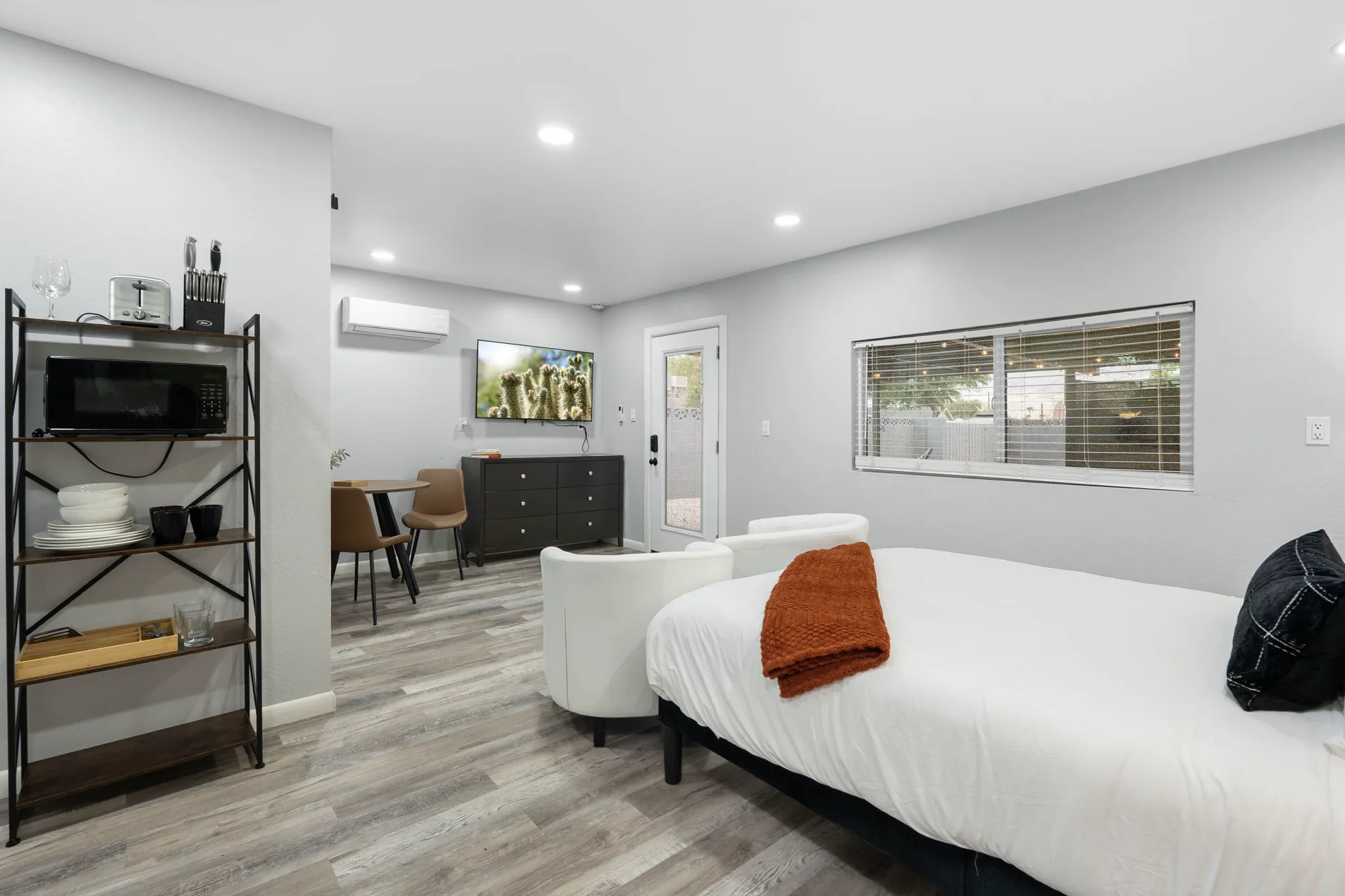 A modern vacation rental studio features a bed, two white armchairs, a small dining area, and a kitchen nook with a microwave and toaster. A TV and window are on the far wall.