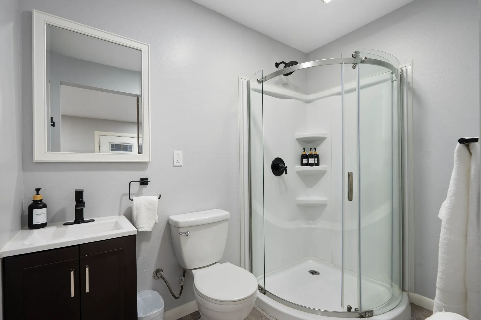 A small, modern bathroom with light gray walls, featuring a corner vanity with a black faucet, a white toilet, and a rounded glass shower with black fixtures.
