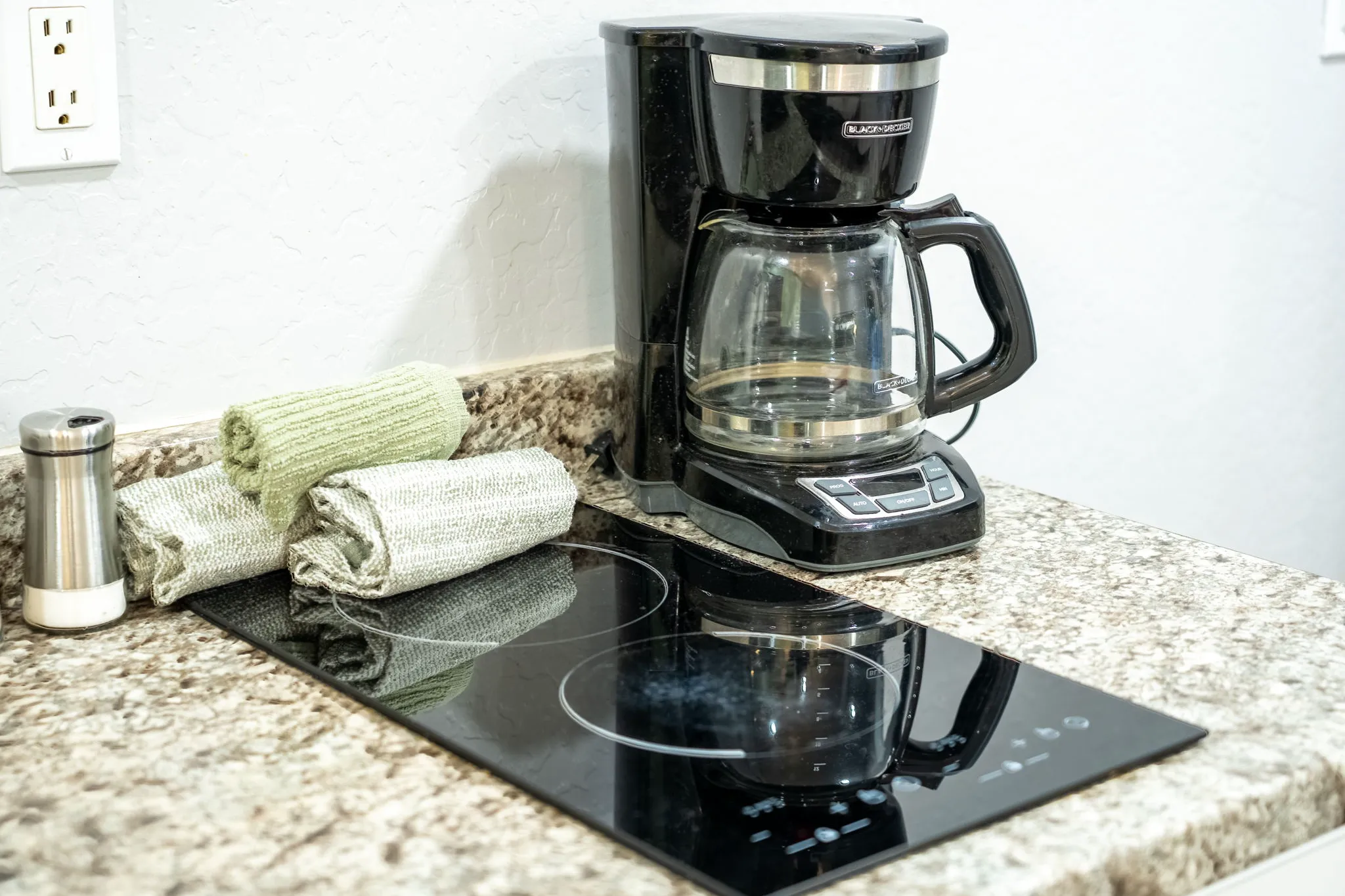 A coffee maker, two rolled hand towels, and a salt shaker are neatly arranged on a granite countertop with a black glass cooktop.