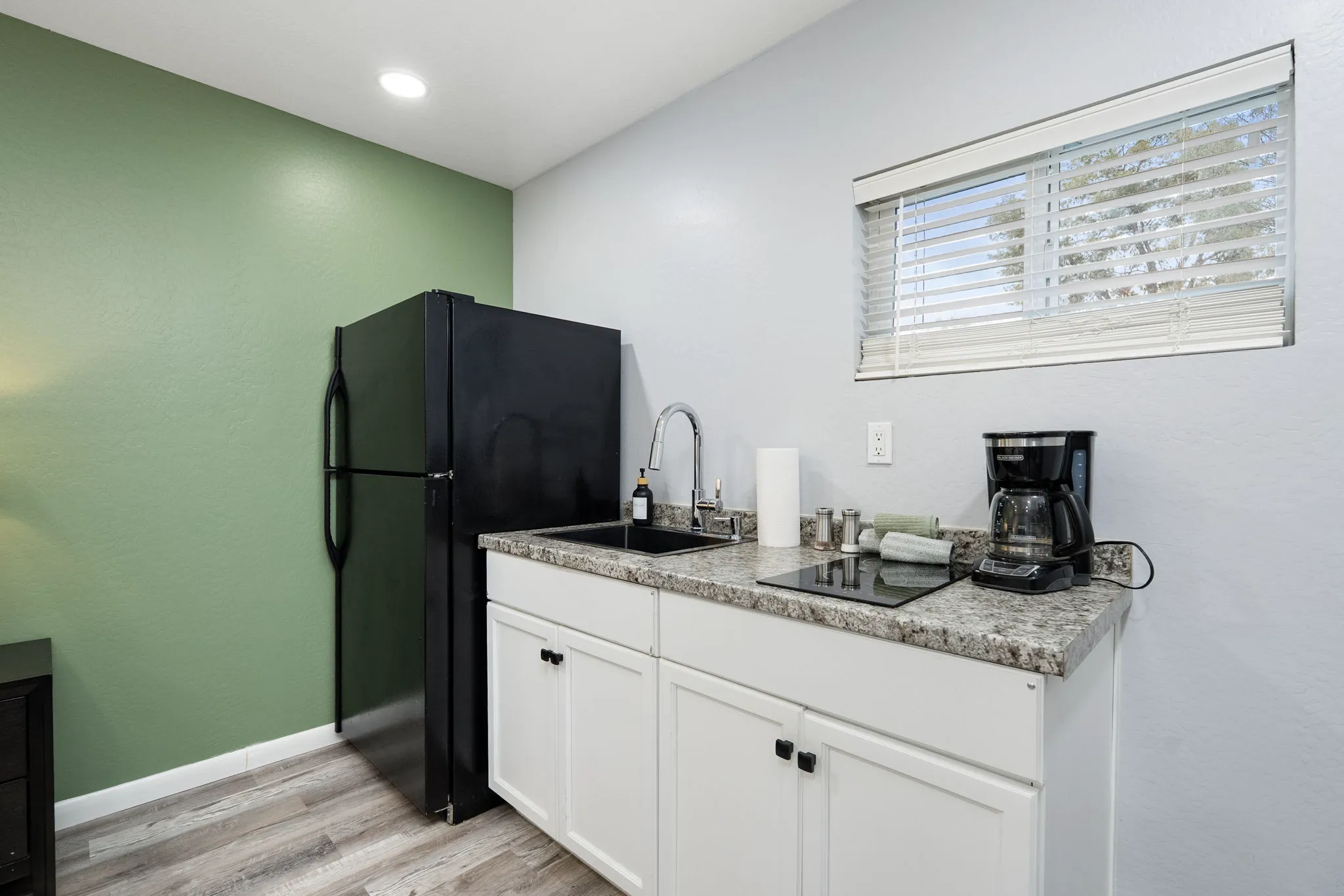 A compact kitchen with a black refrigerator, white cabinets, speckled gray countertops, a sink, induction cooktop, and coffee maker, against a green accent wall.