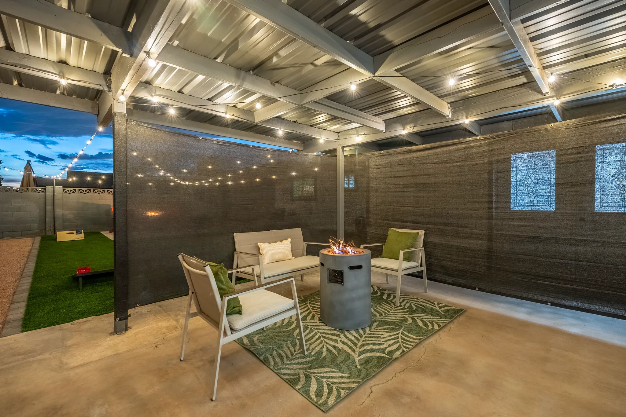 An outdoor patio at dusk with string lights illuminating a seating area around a lit fire pit. It includes two chairs, a loveseat on a green rug, and a cornhole game nearby.