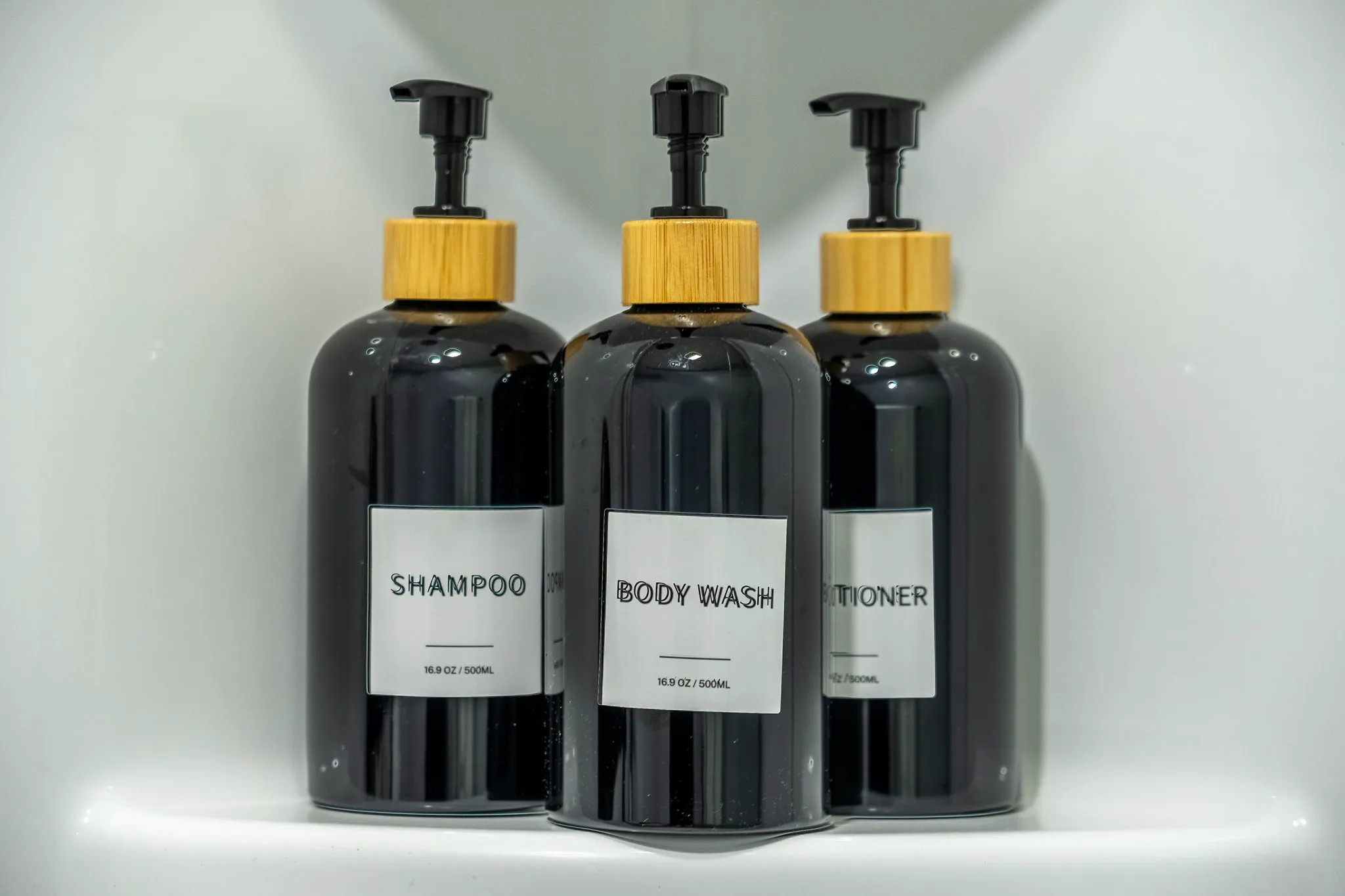 Three black pump bottles with wooden tops and white labels reading 'SHAMPOO,' 'BODY WASH,' and 'CONDITIONER' are neatly arranged on a white surface.