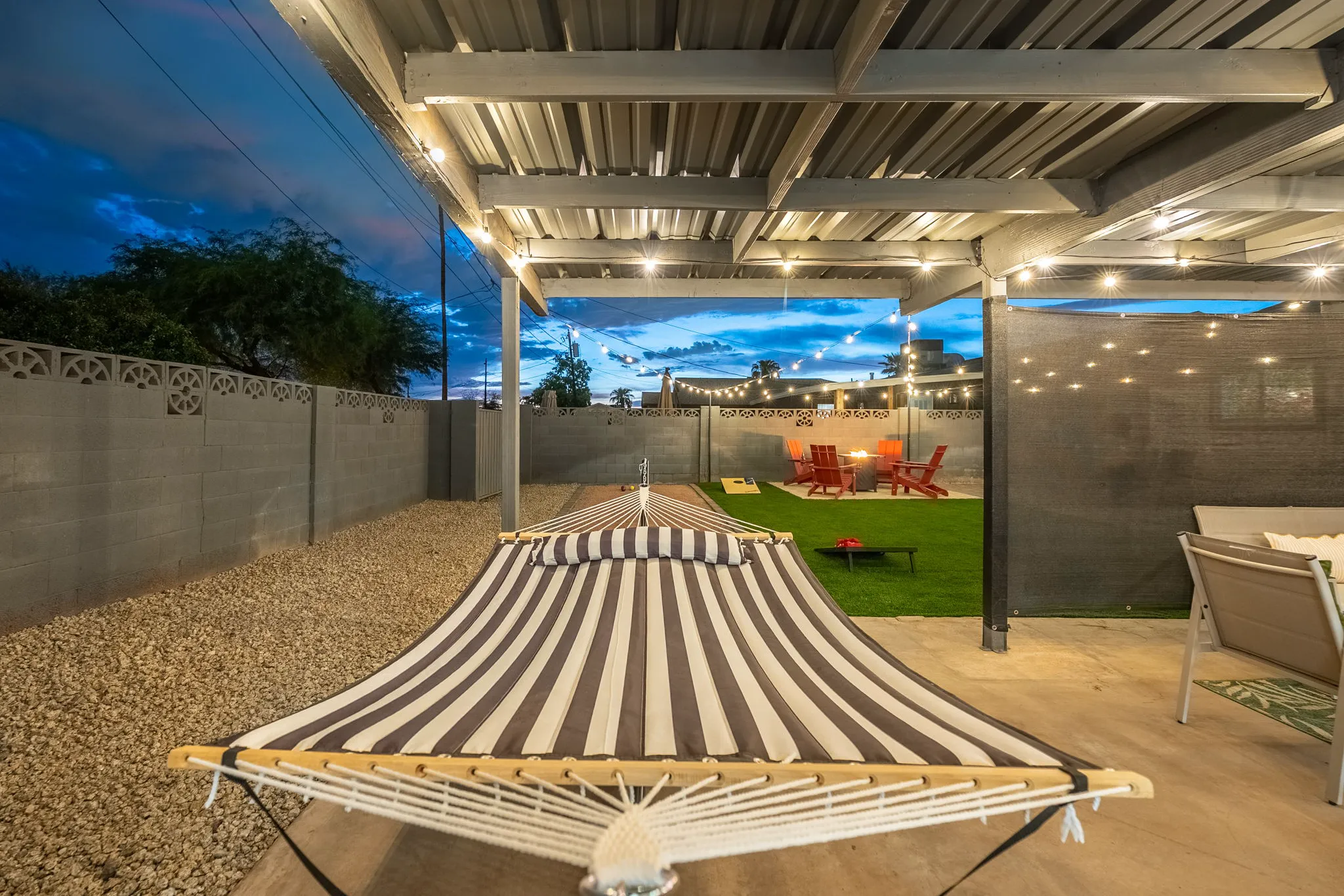 A striped hammock under a covered patio with string lights, looking out to a backyard featuring artificial turf, a fire pit, and yard games at twilight.