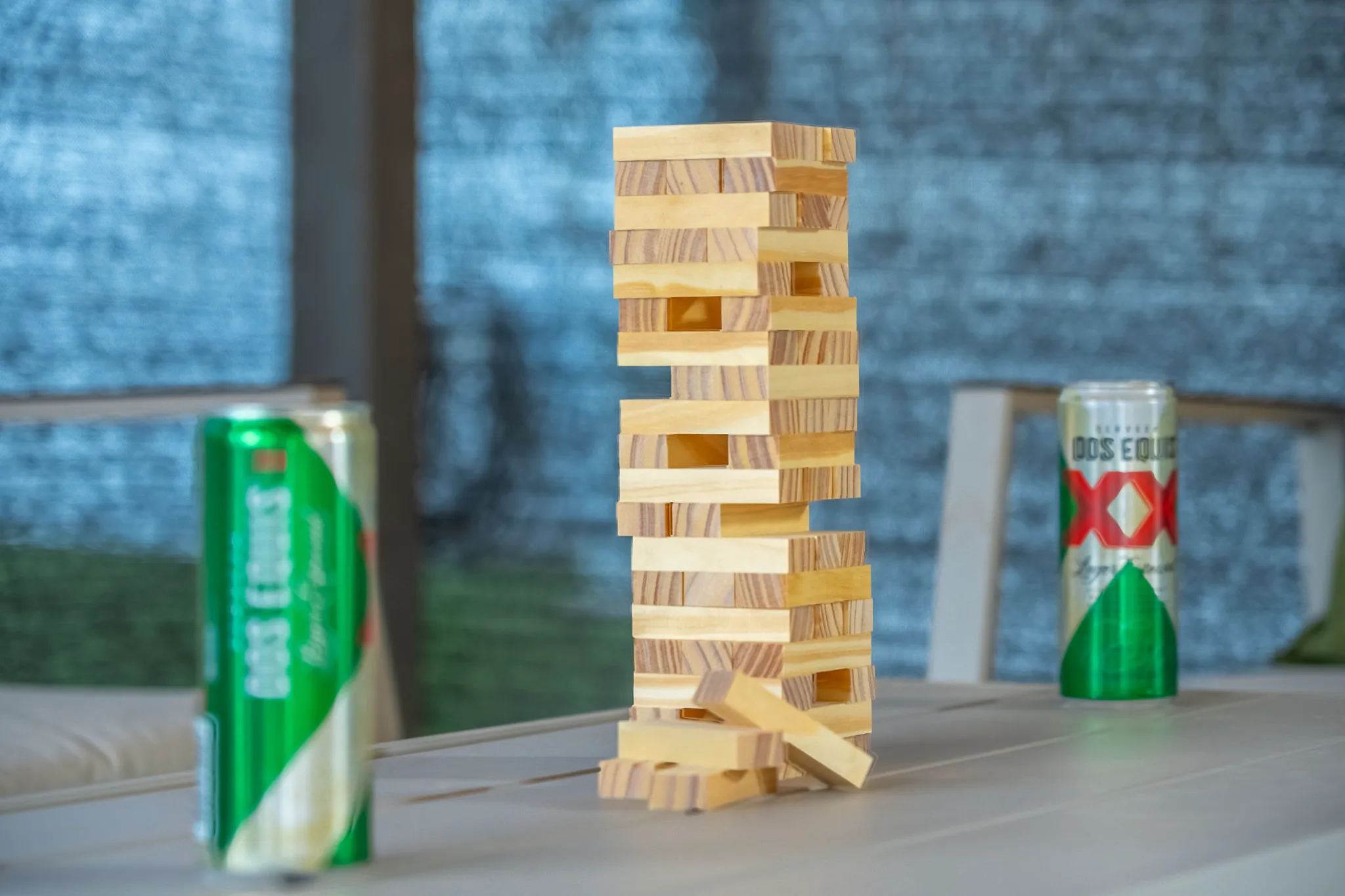 A Jenga tower stands precariously on a table, with several blocks removed and lying nearby. Two Dos Equis beer cans are on either side, against a blurred blue background.
