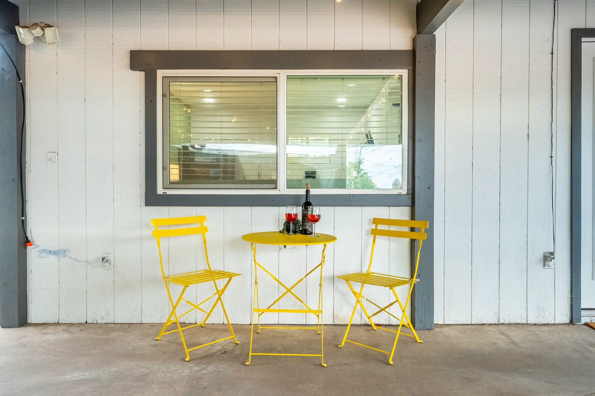 An outdoor patio features a yellow round table with a wine bottle and two glasses, flanked by two yellow chairs, set against a white wall with a window.