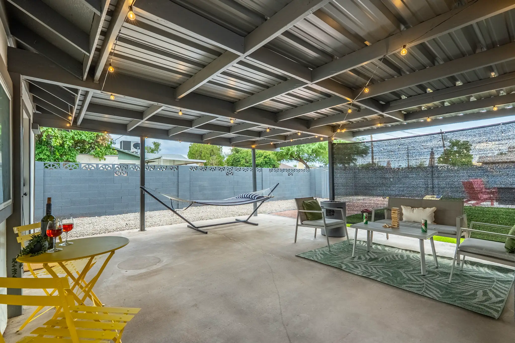A covered outdoor patio features a yellow bistro set with wine, a striped hammock, and a lounge area with gray furniture on a green patterned rug, under a string-light adorned roof.