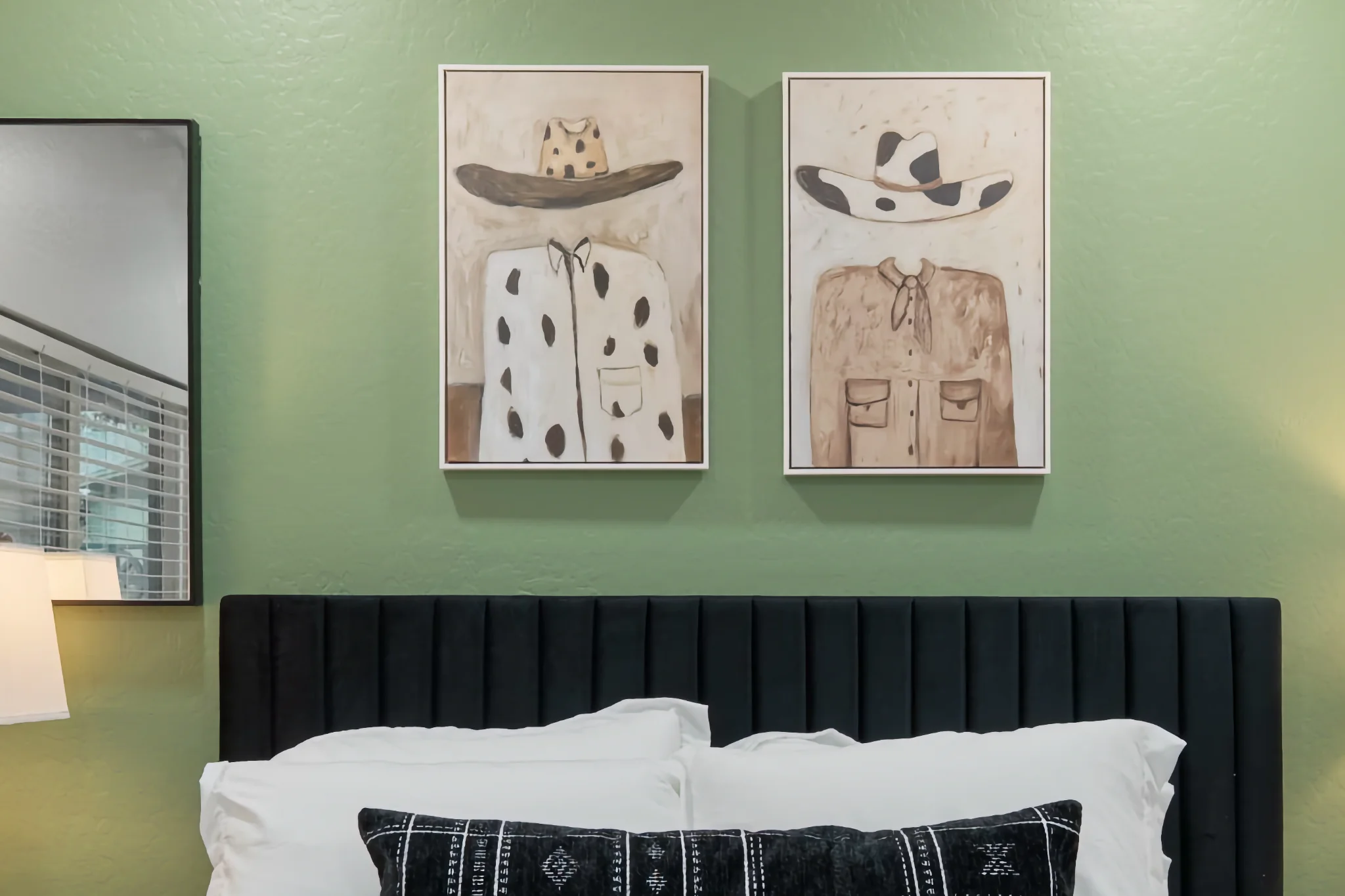A bedroom with a green textured wall, a black padded headboard, and white bedding. Two framed artworks depicting cowboy hats and shirts hang above the bed.