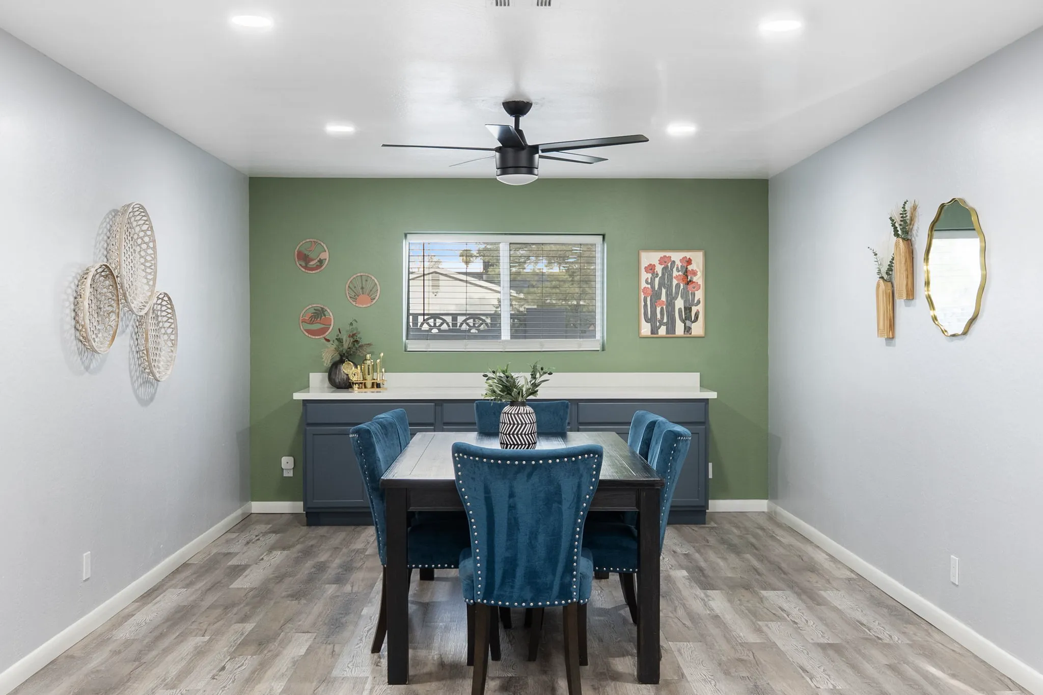 A modern dining room with a dark wood table and six blue velvet chairs. A sage green accent wall has a window, white counters, and wall art, while side walls are light grey.