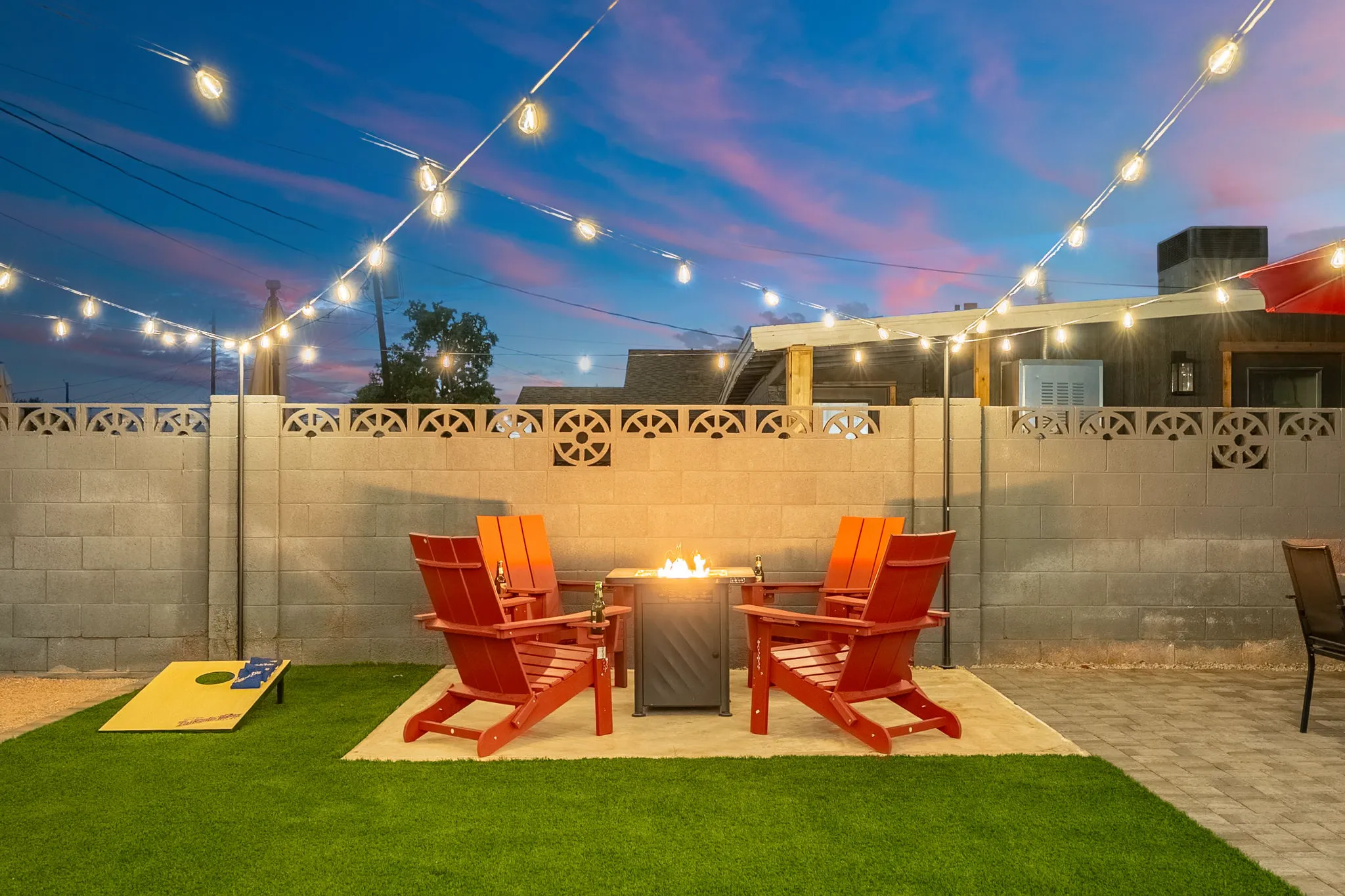 An outdoor patio at dusk has a lit fire pit, four red Adirondack chairs, and string lights. Artificial grass includes a yellow cornhole game under a colorful sunset sky.