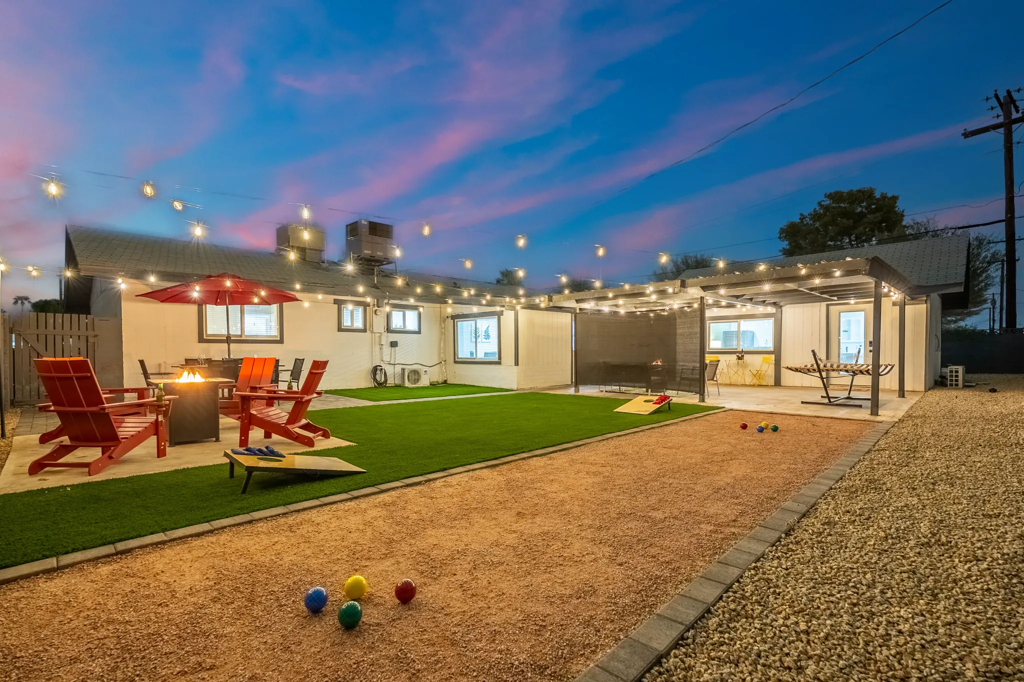 A vibrant backyard at dusk featuring a bocce ball court, artificial turf, a fire pit with red chairs, string lights, and a covered patio with a hammock.