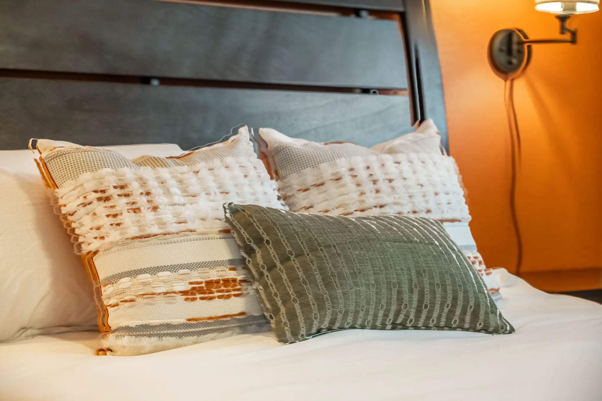 Three decorative pillows—two textured white and orange, one green striped—are arranged on a white bedspread in front of a dark wooden headboard. A wall lamp illuminates an orange wall.