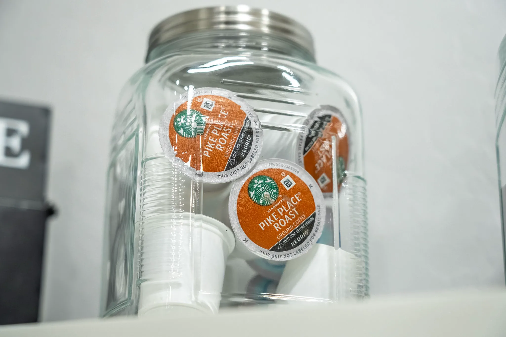 A clear glass jar filled with orange Starbucks Pike Place Roast Keurig K-Cup pods and small white plastic cups sits on a counter.