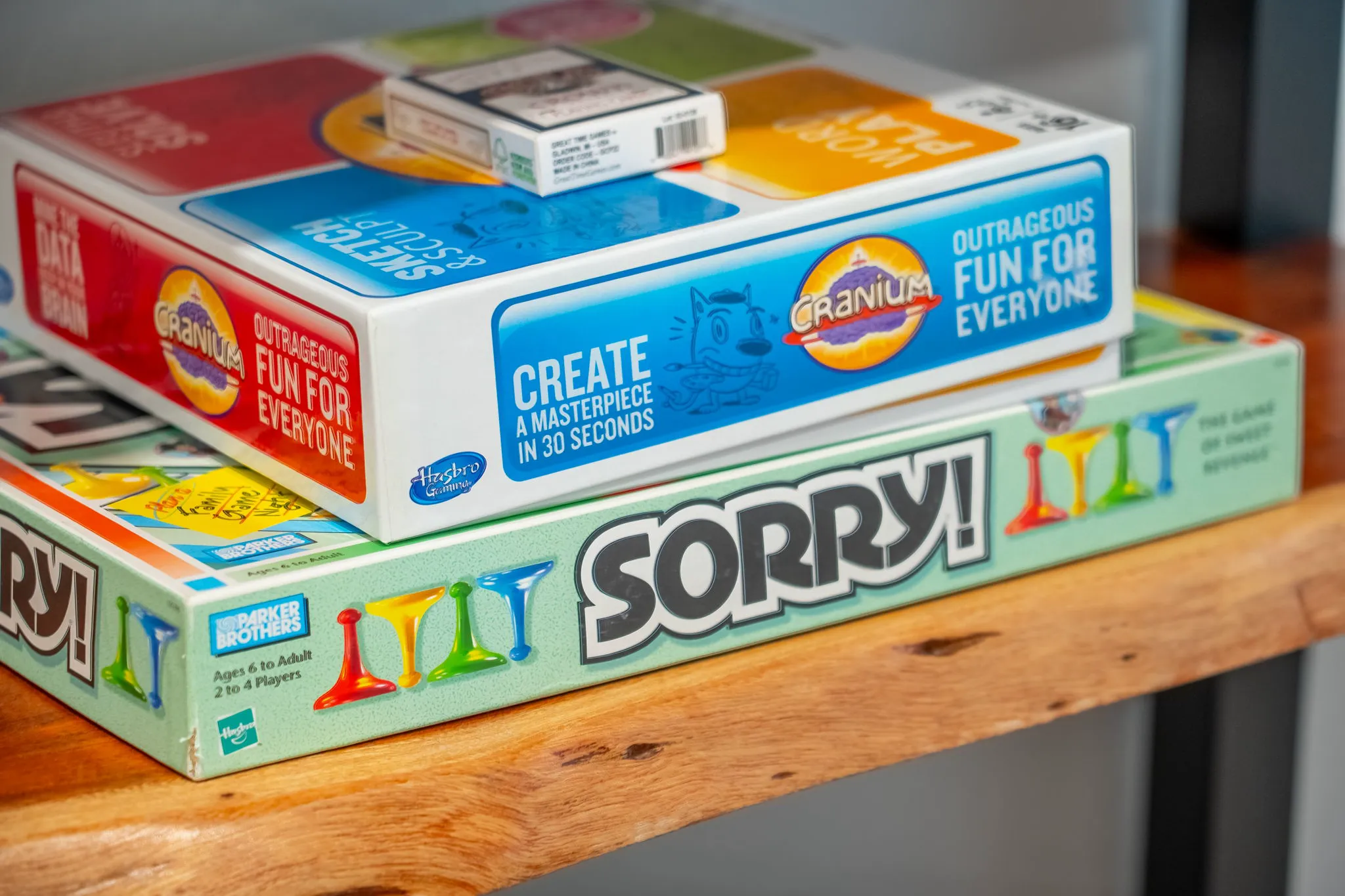 Two colorful board game boxes, Cranium and Sorry!, are stacked on a natural wood shelf, with a small box of cards on top.