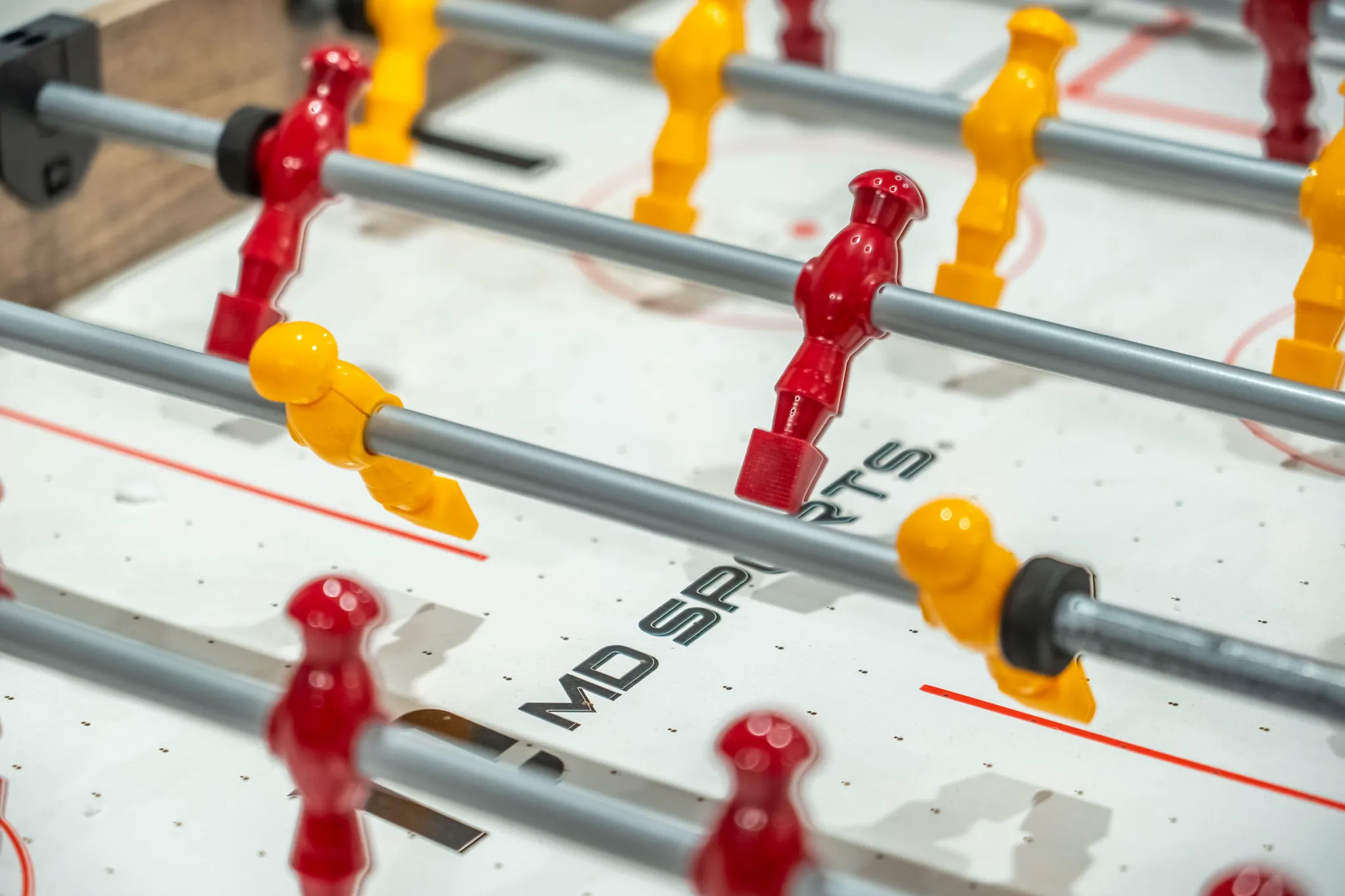 A close-up view of a foosball table showing red and yellow plastic players on metal rods, with the "MD SPORTS" logo visible on the white playing surface.