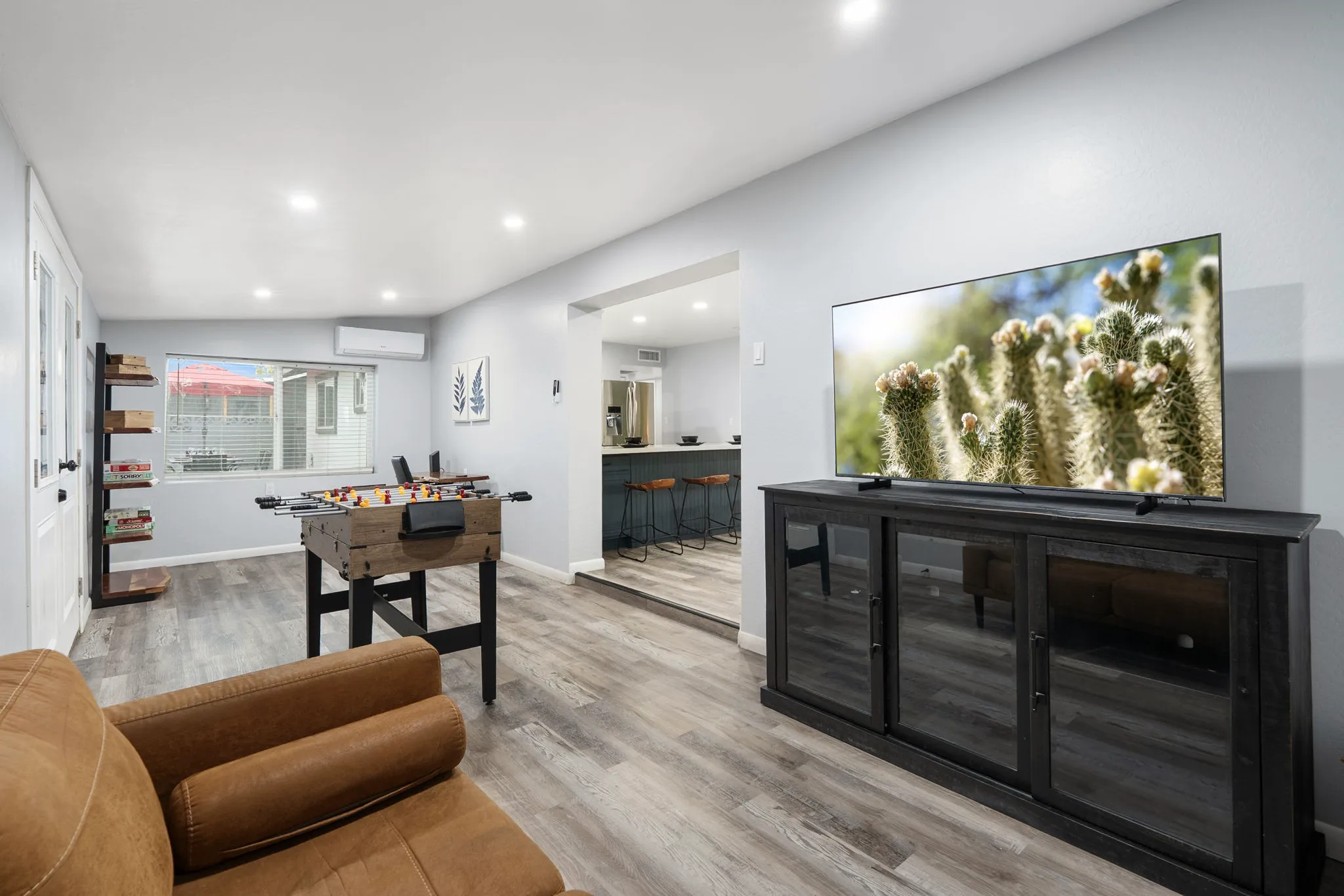 A modern living room features a foosball table, a large TV displaying a cactus, and a brown leather couch. The room has light wood-look flooring and white walls, with an adjacent kitchen visible.