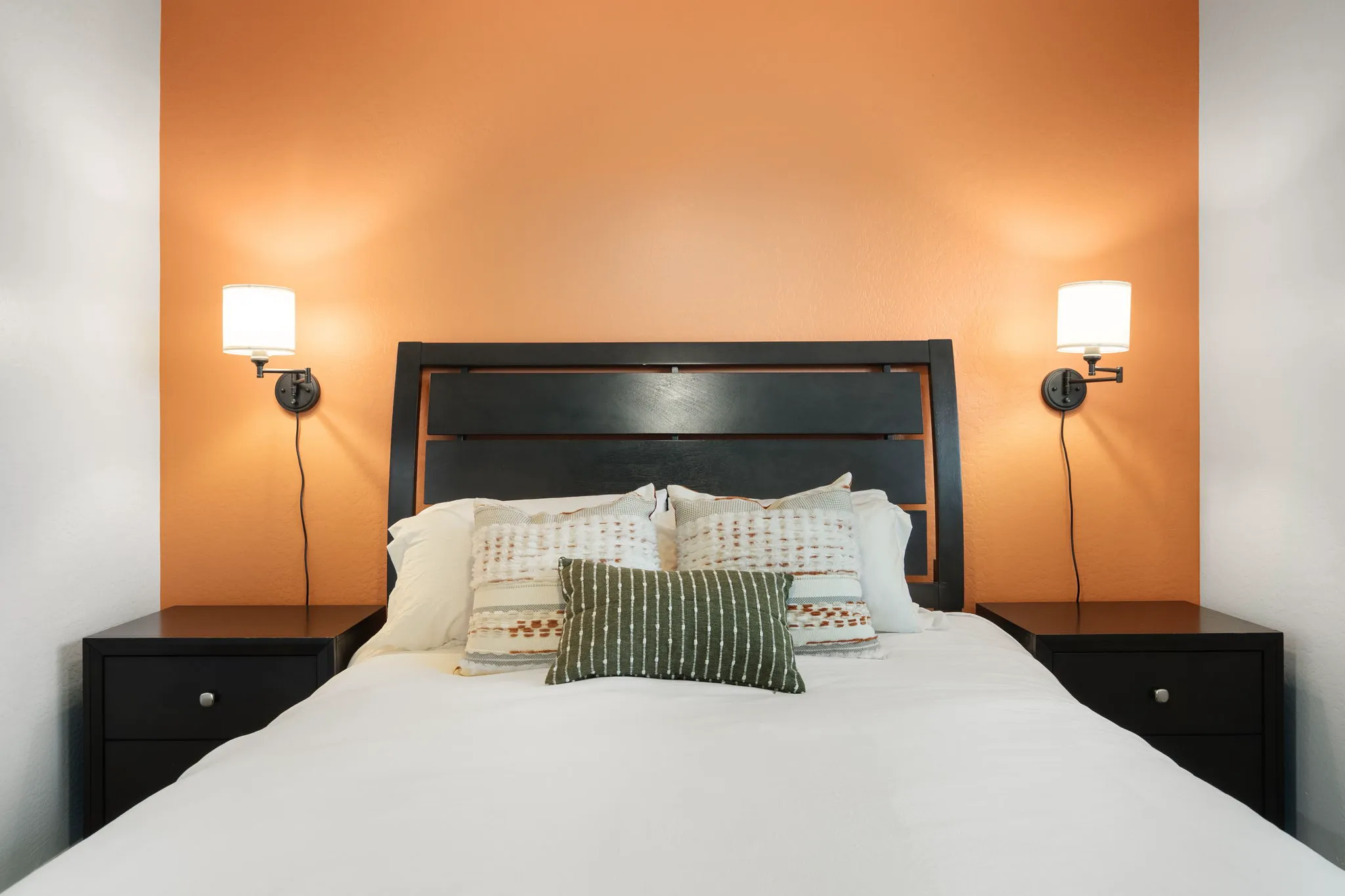 A bedroom features a bed with white sheets and decorative pillows, flanked by two dark nightstands. An orange accent wall is behind the dark wooden headboard, with two wall sconces.