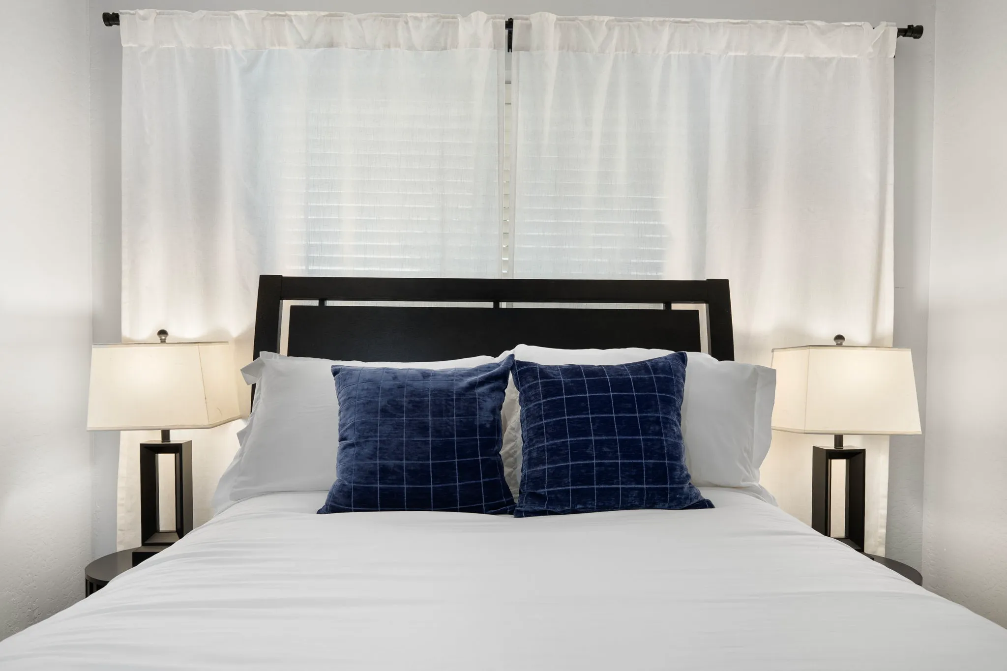 A neatly made bed with white sheets and two blue patterned pillows. A dark headboard is behind it, flanked by two nightstands with lamps and white curtains on the windows.