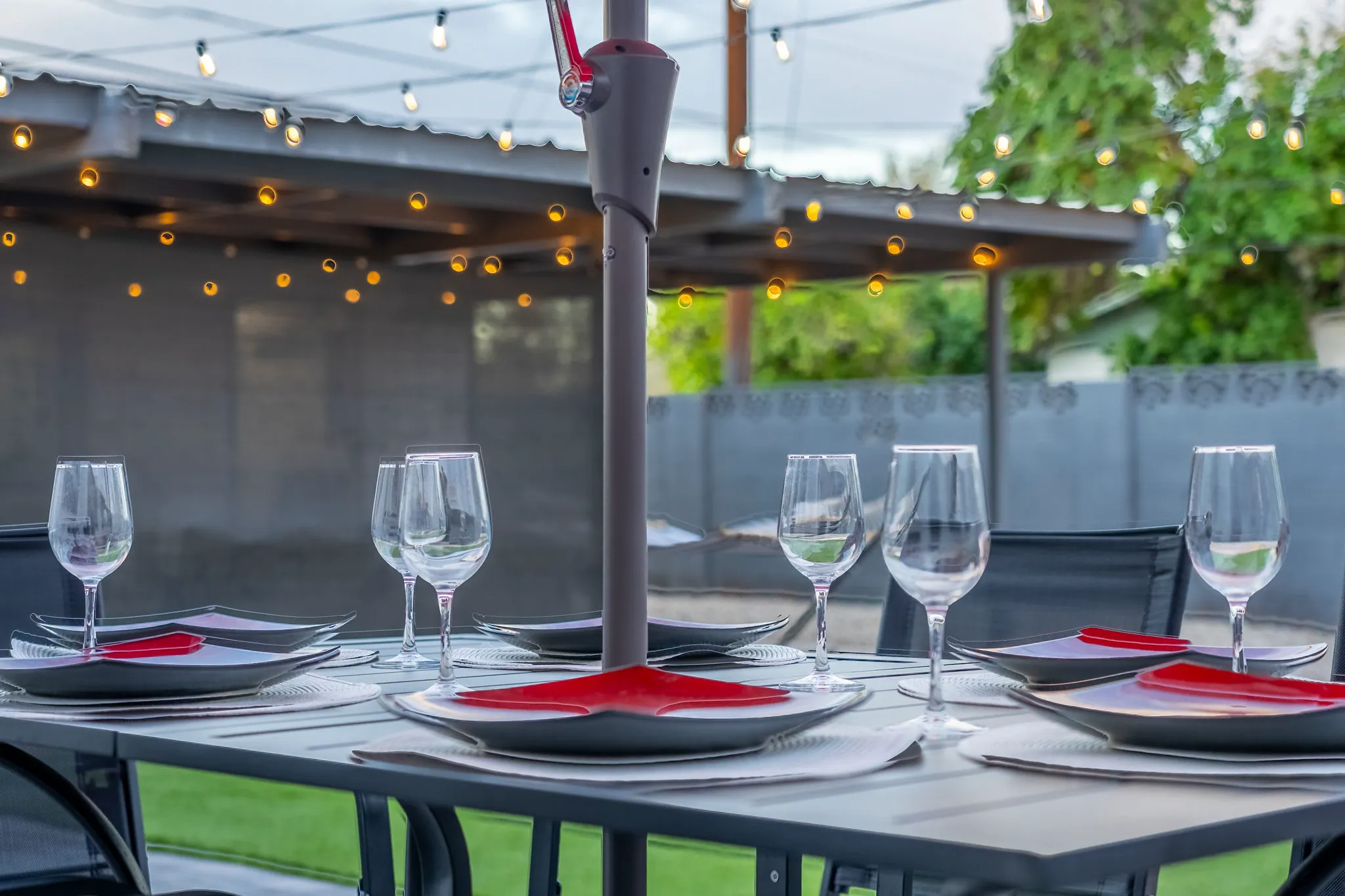An outdoor patio dining table set for guests with black and red plates, wine glasses, and an umbrella post in the center, under string lights.
