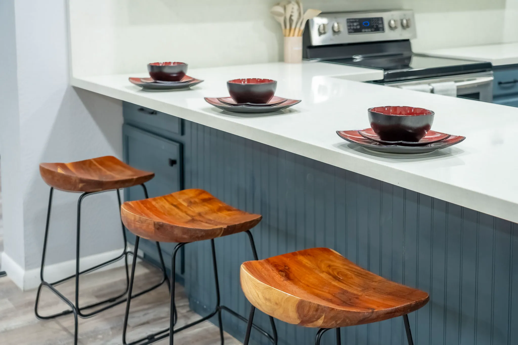 A kitchen island with a white countertop and three wooden bar stools. Three place settings of black and red dishes are arranged on the counter, which has dark blue beadboard paneling.
