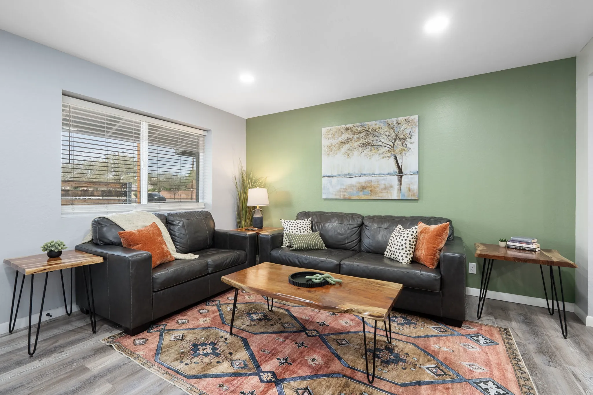 A cozy living room features two dark leather sofas with colorful pillows, a natural wood coffee table on a patterned rug, and a green accent wall with tree art.