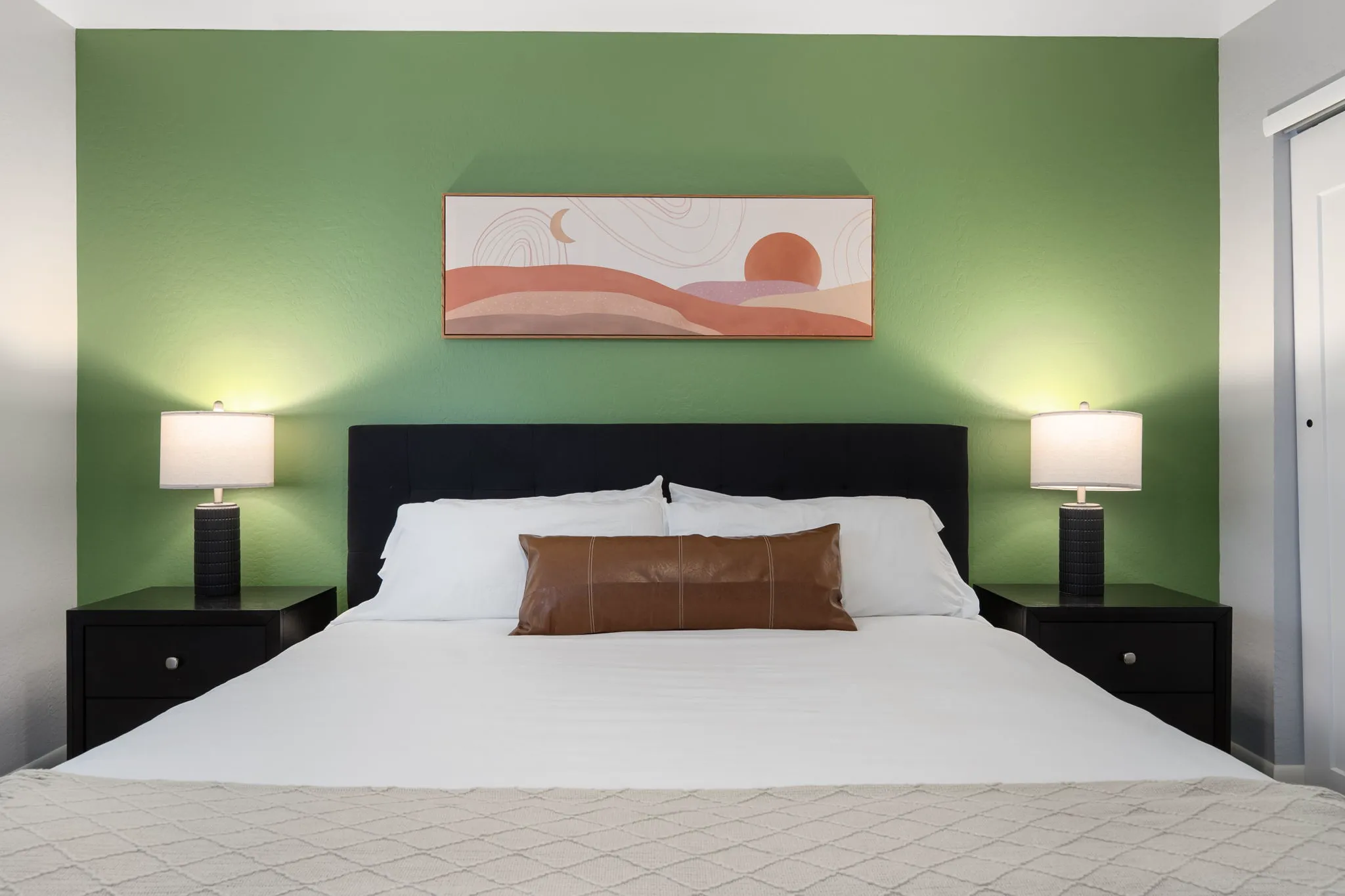 A bedroom features a bed with white linens, a brown pillow, and a cream blanket against a green accent wall. Black nightstands with illuminated lamps flank the bed, with abstract art above.