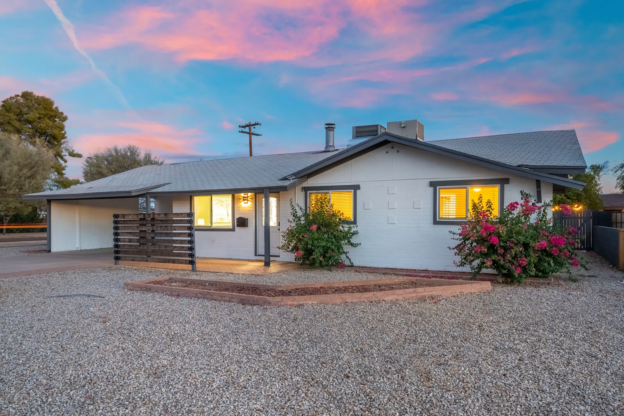 A single-story white house with gray trim, illuminated windows, and a carport sits under a vibrant sunset sky. The gravel front yard features flowering bushes.
