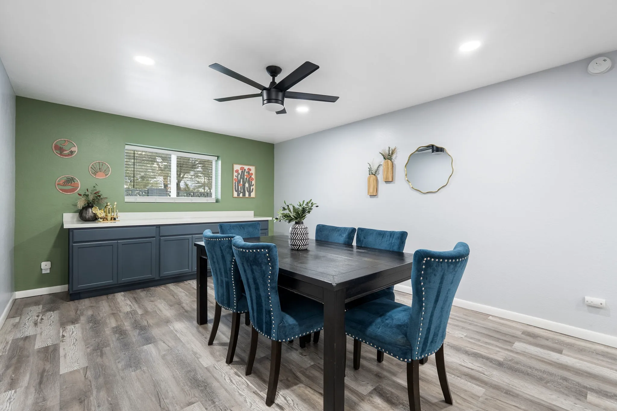 A modern dining room features a dark wooden table with six teal studded chairs, a ceiling fan, a window, and a built-in cabinet against a green accent wall.