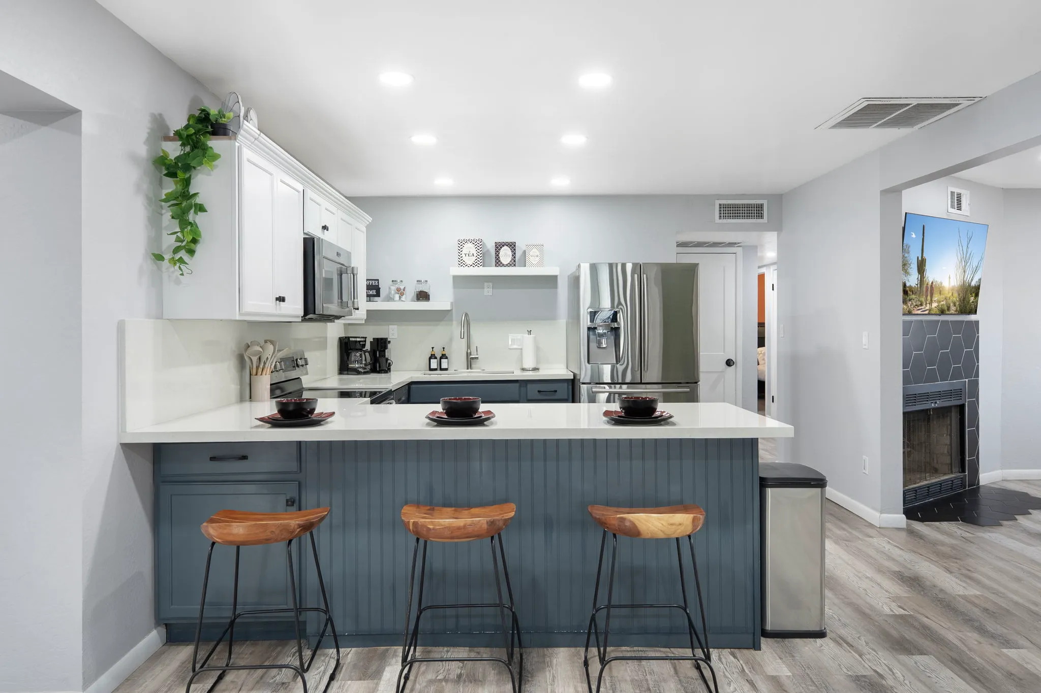 A modern kitchen with white upper cabinets, dark blue lower cabinets, and a kitchen island with three wooden bar stools. Stainless steel appliances and light wood floors create a clean space.