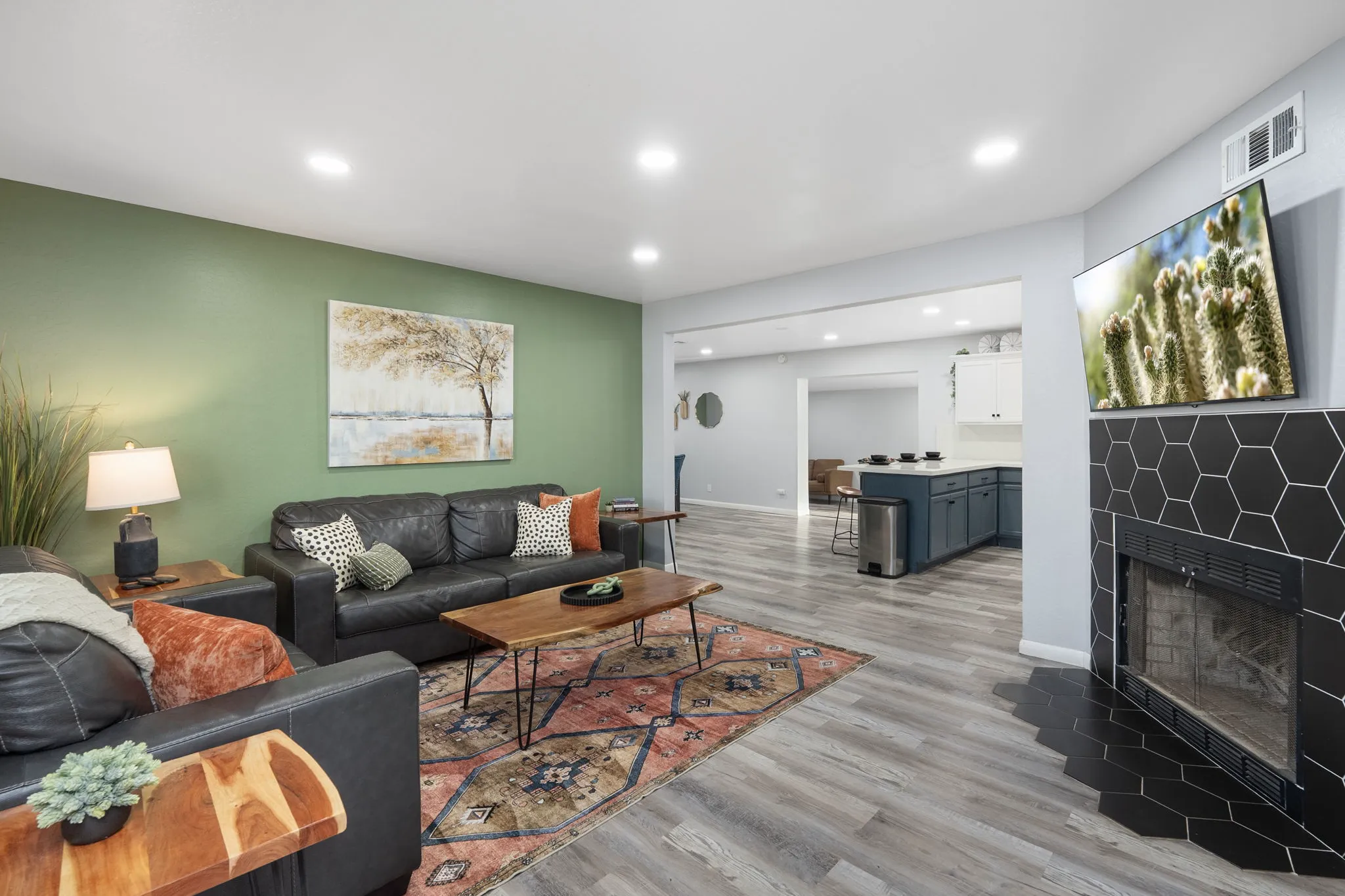 A modern living room has dark leather sofas, a patterned rug, wooden coffee table, green accent wall, and a black hexagonal-tiled fireplace with a TV. A kitchen is visible.