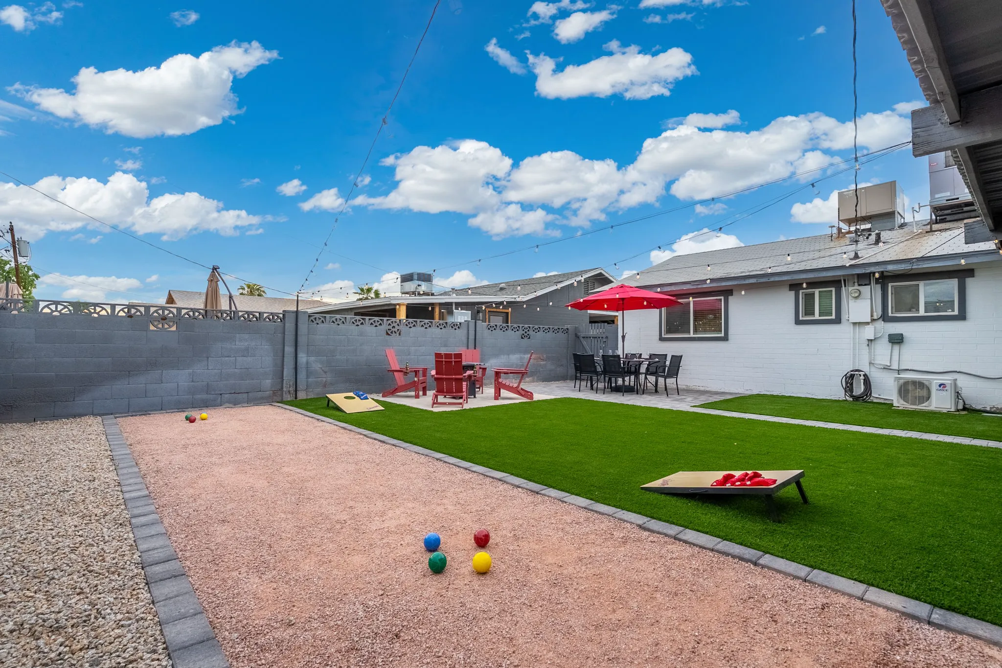 A sunny backyard with a bocce ball court, artificial turf, and a patio featuring red Adirondack chairs, a fire pit, and a dining set under a red umbrella.