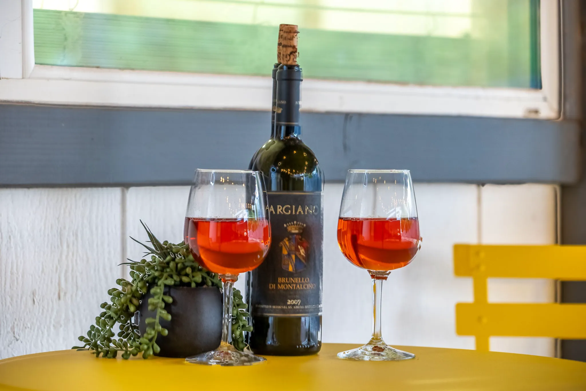 A bottle of Argiano Brunello di Montalcino 2007 and two glasses of rosé wine sit on a yellow table with a small plant. A yellow chair is in the background.