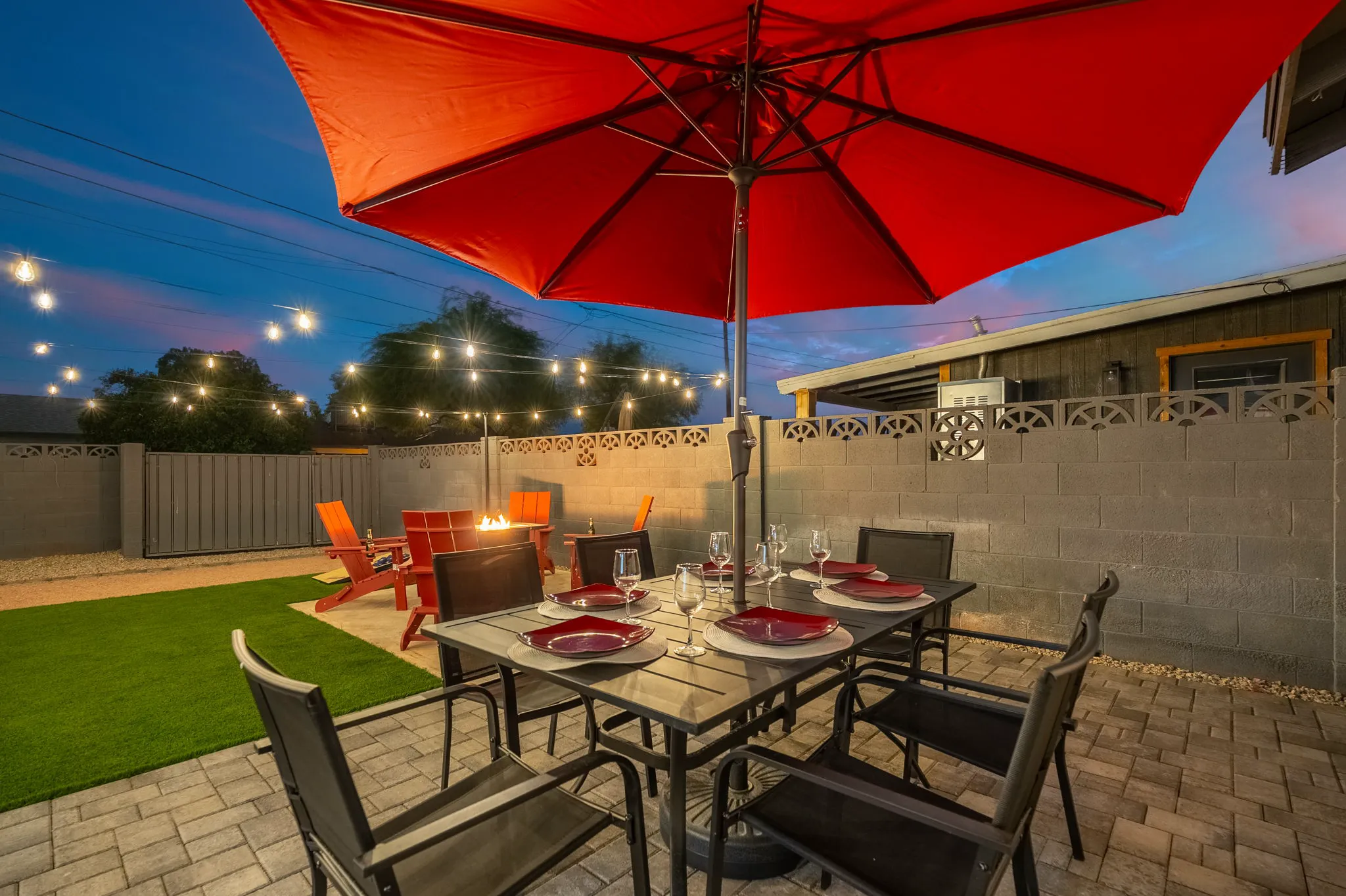 A twilight backyard patio with a dining table for six under a red umbrella. String lights illuminate a fire pit, red chairs, artificial grass, and paved areas.
